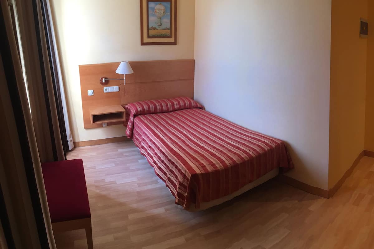 Habitación de hotel con una cama, un escritorio con computadora y controles remotos, una silla, una lámpara y una puerta.
