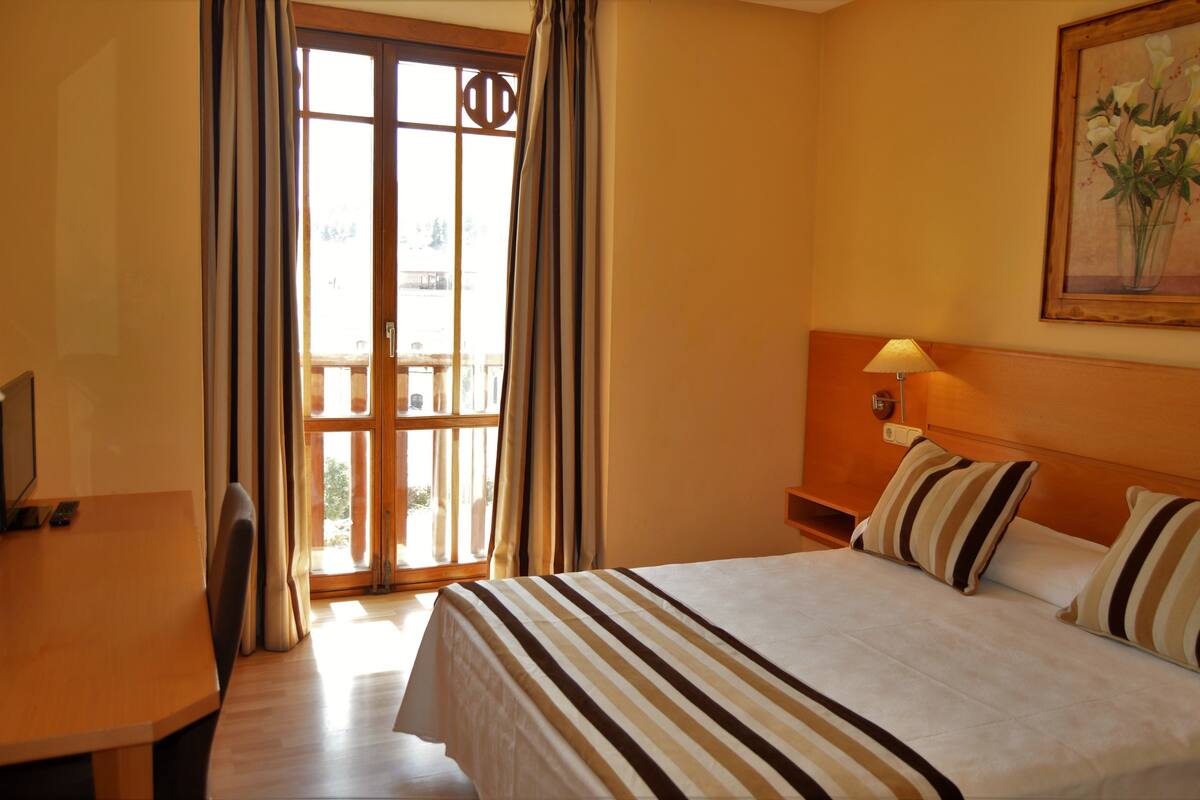 Habitación de hotel con cama, escritorio, silla, ventana con cortinas y un cuadro en la pared.