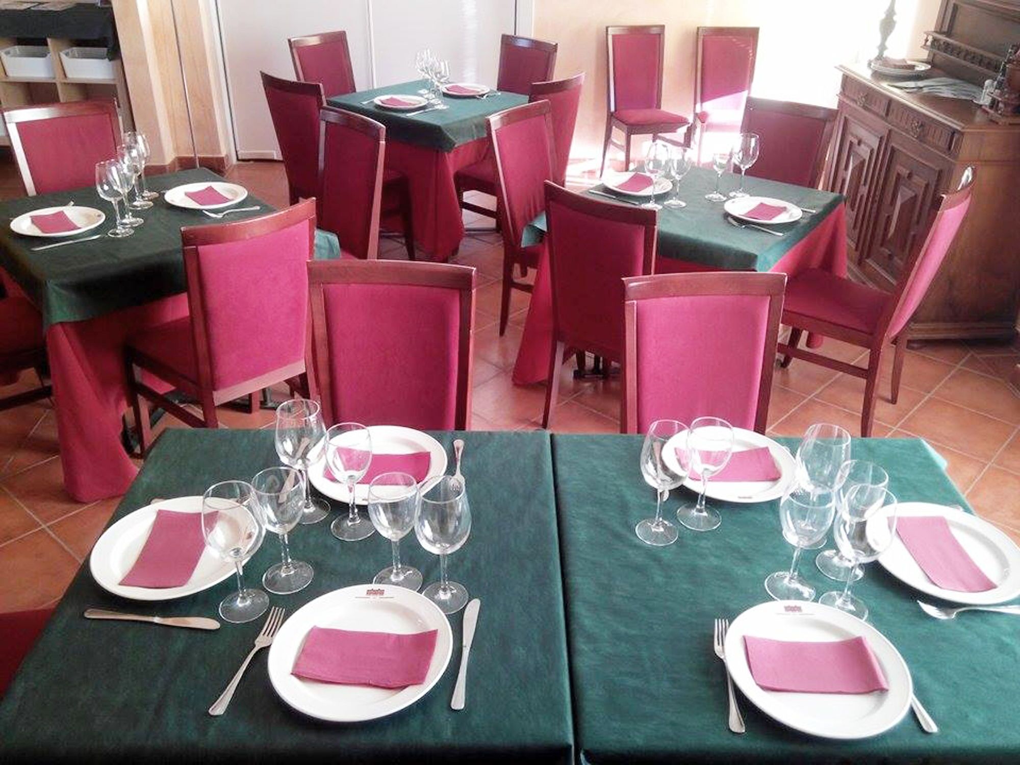 Zona de menjador amb taules preparades per a quatre, cadascuna amb mantells verds, caires vermells i plats blancs amb serpentines rosa.