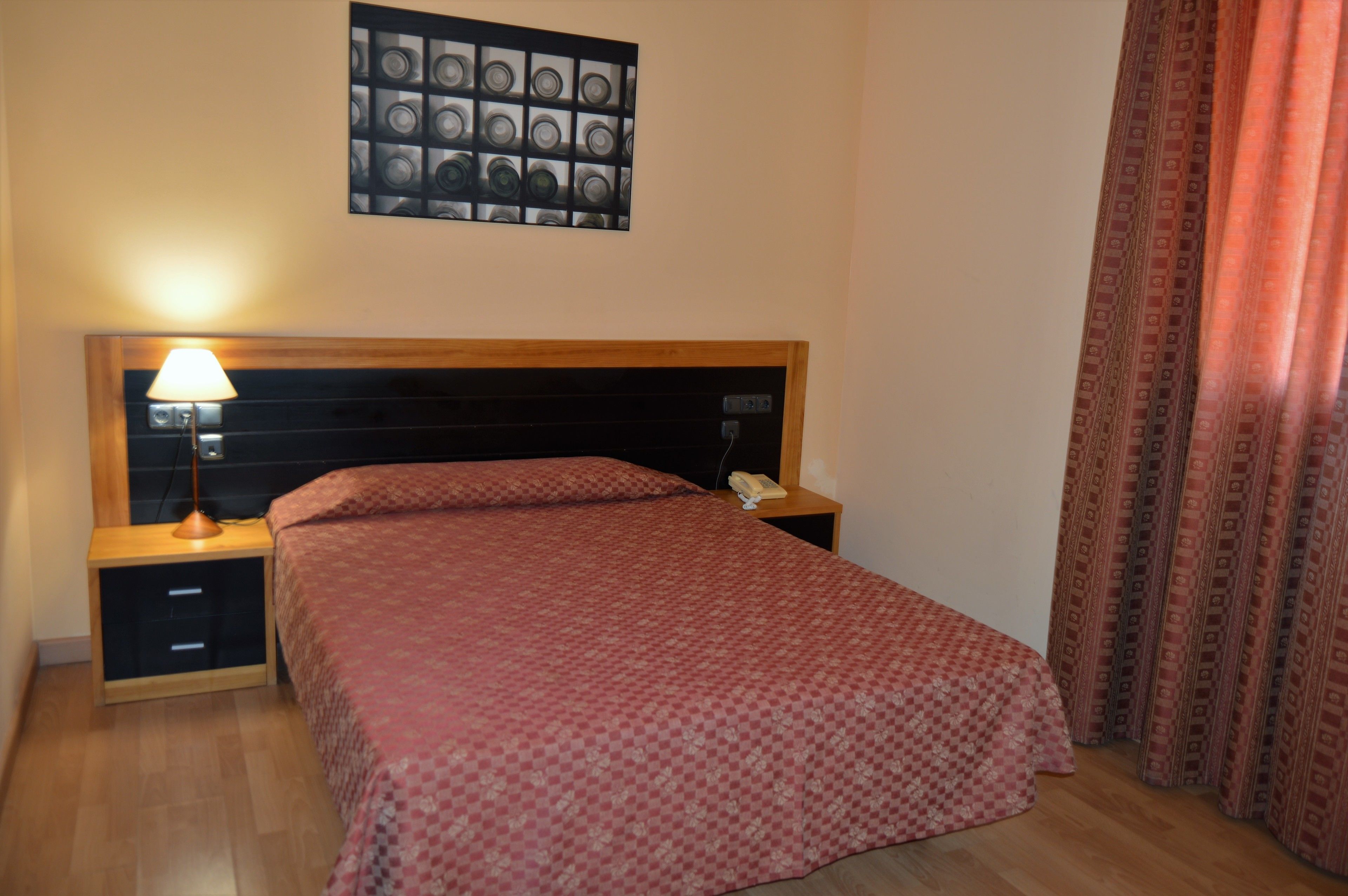 Una cama bien hecha con una colcha estampada, una lámpara de noche, una mesita de noche con un teléfono y una obra de arte en la pared sobre la cama.