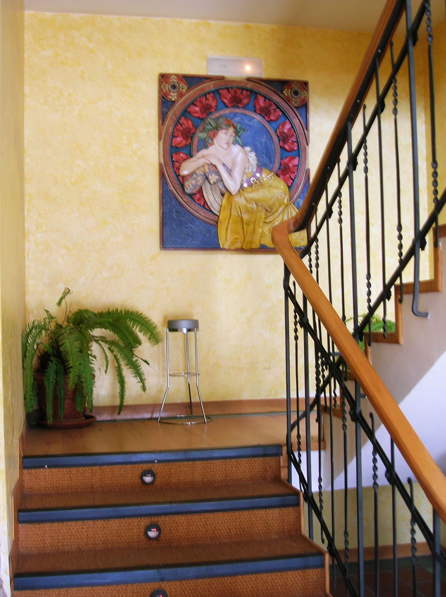 A staircase with a wooden handrail and black balusters, a potted fern, a metal stool, and a decorative painting on the wall.