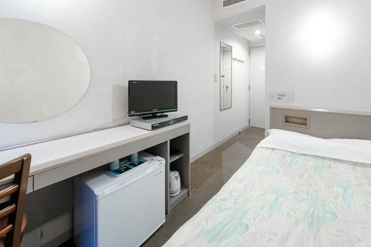 A hotel room with a bed, a desk with a computer, a chair, a small trash bin, and a mirror.