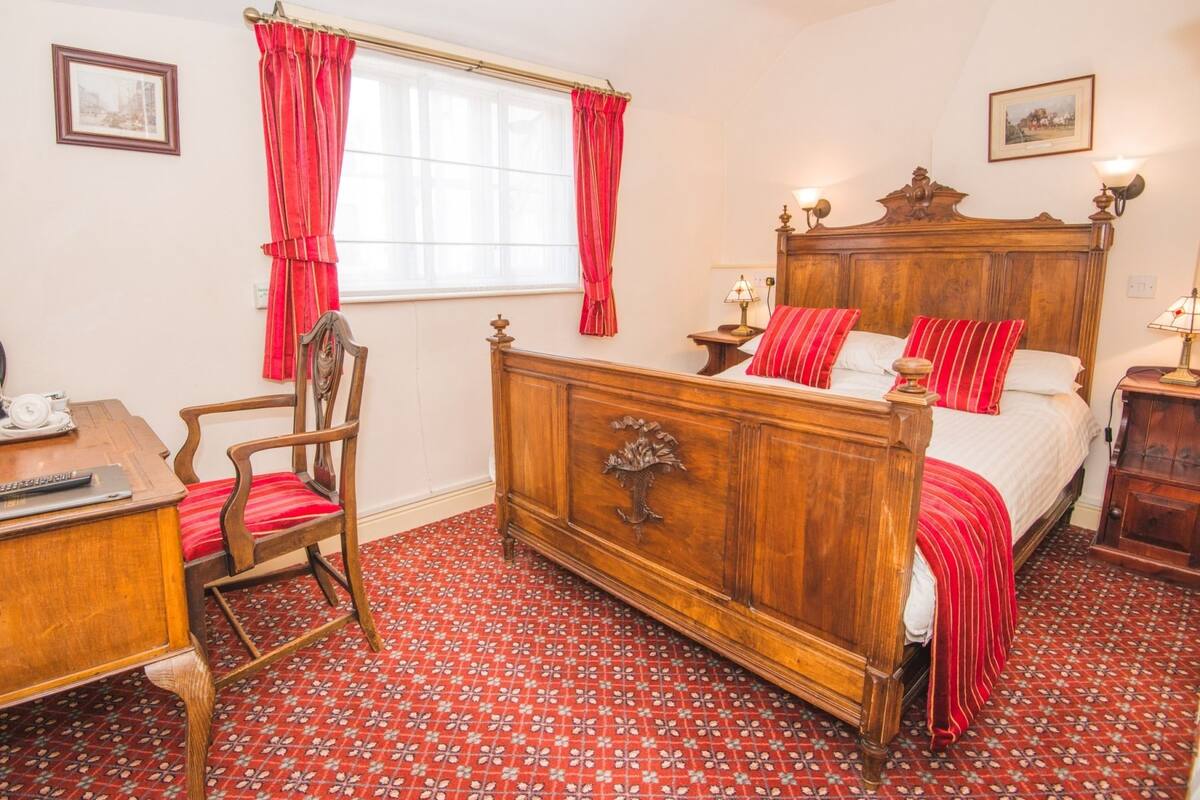 A bedroom with a wooden bed, red curtains, a desk with a chair, and a patterned carpet.