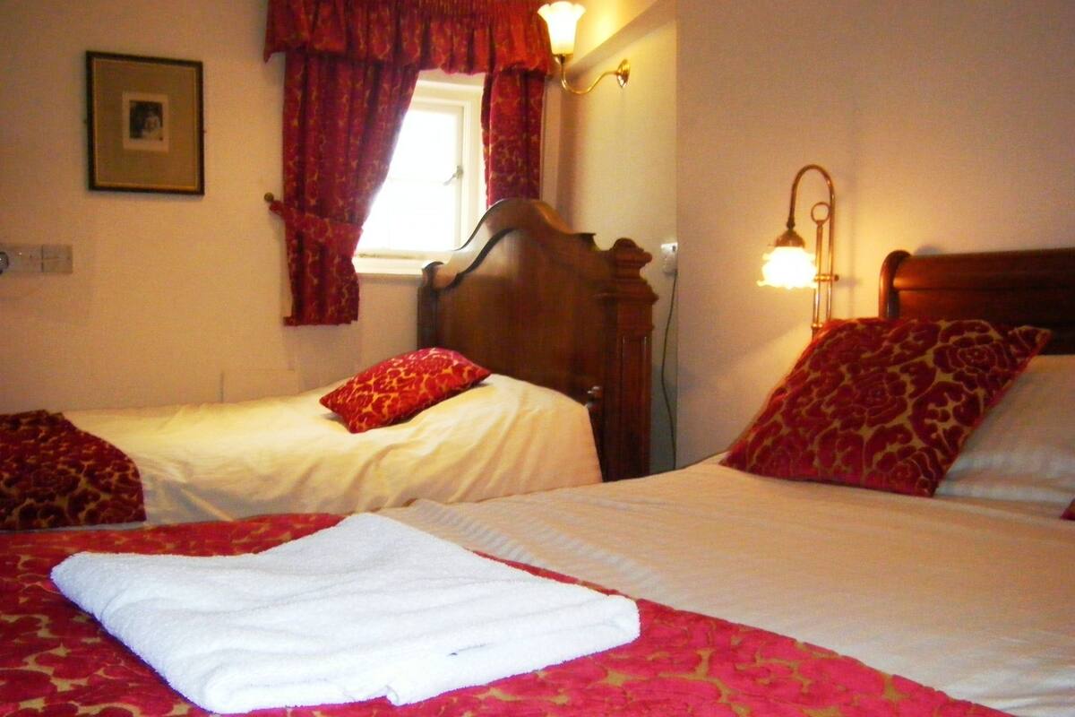 A hotel room with two beds, red and white bedding, a window with red curtains, and a wall-mounted lamp.