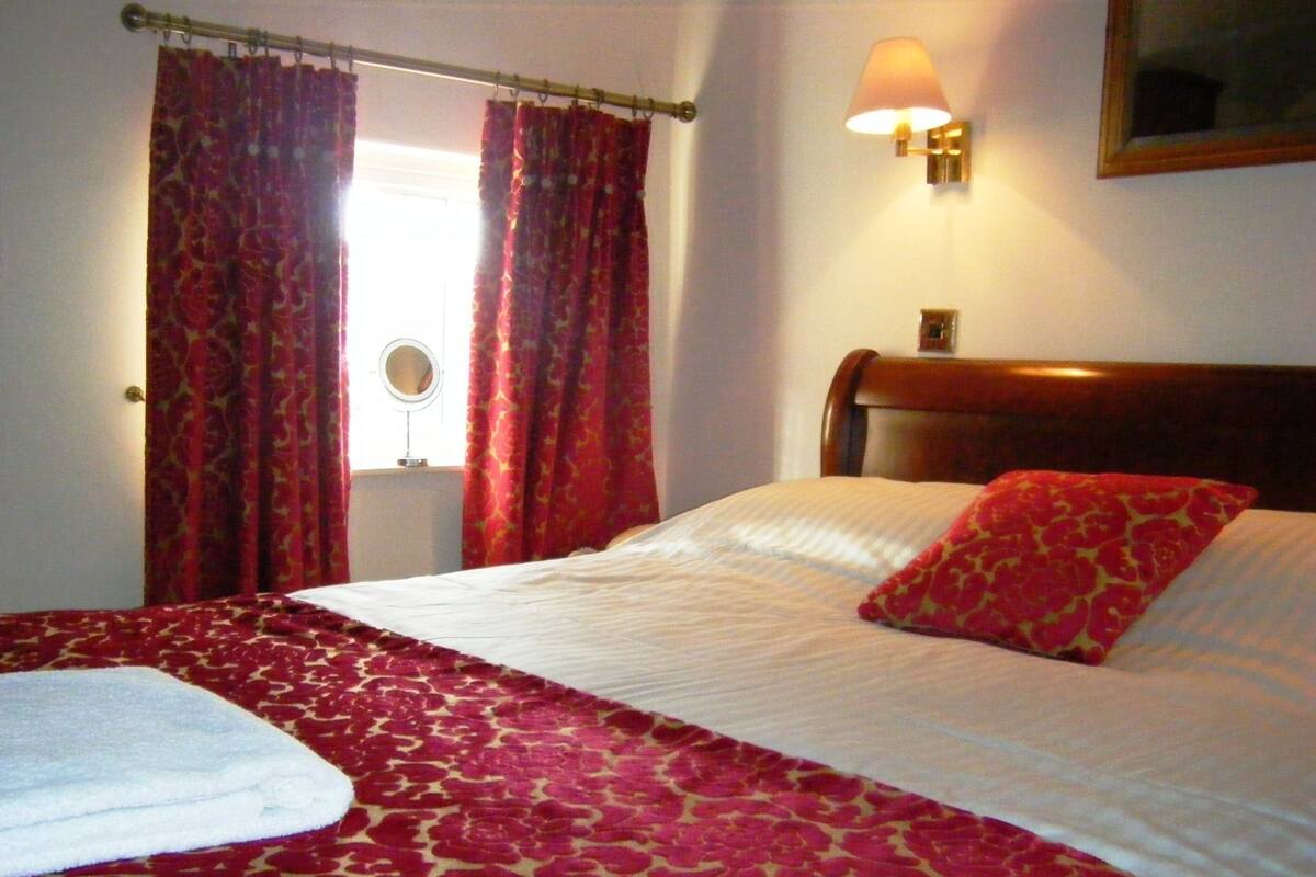 A neatly made bed with a red and white patterned bedspread, a single red pillow, and a wall-mounted lamp.