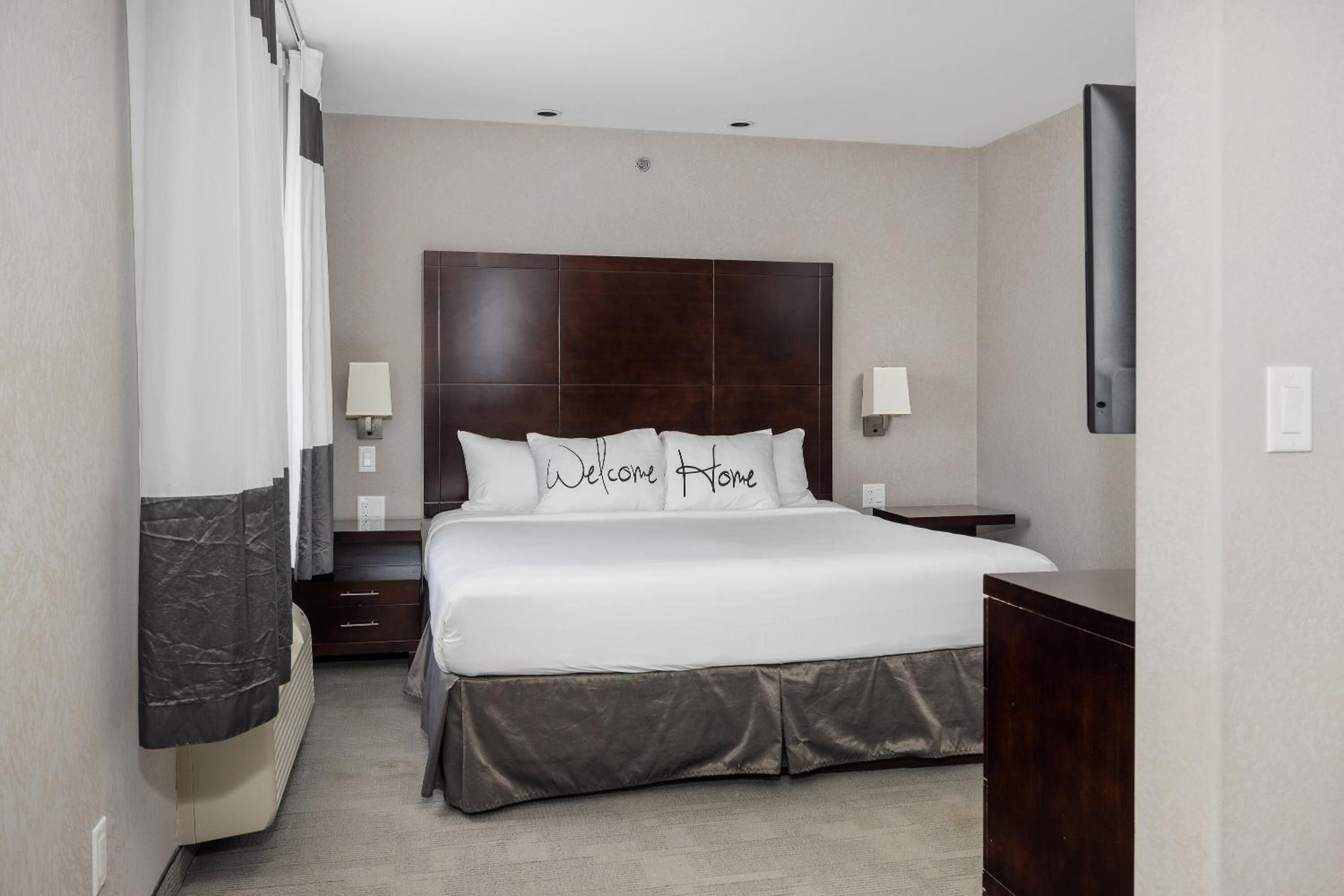 A hotel room with a large bed, two bedside tables, a flat-screen TV, and a dark wooden headboard.