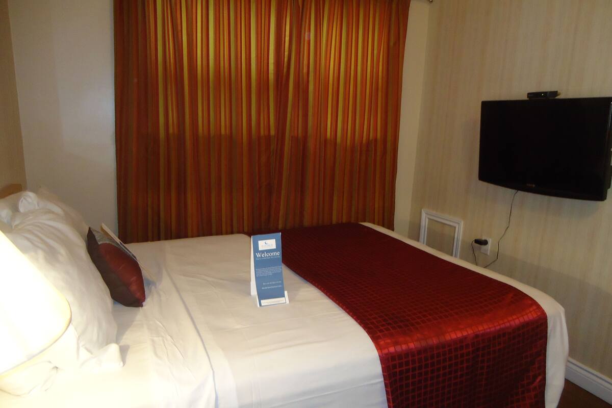 Une chambre d’hôtel avec un lit, un panneau d’accueil, un téléviseur fixé au mur et un rideau rouge.