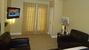 A living room with a ceiling fan, a television mounted on the wall, a sofa, a small table with a lamp, and a door with curtains. - Bayside Inn & Waterfront Suites (Kingston)