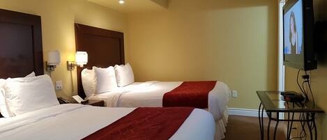 A hotel room with two beds, each with white linens and red bedspreads, a nightstand with a lamp, a TV mounted on the wall, and a small table with a remote control.