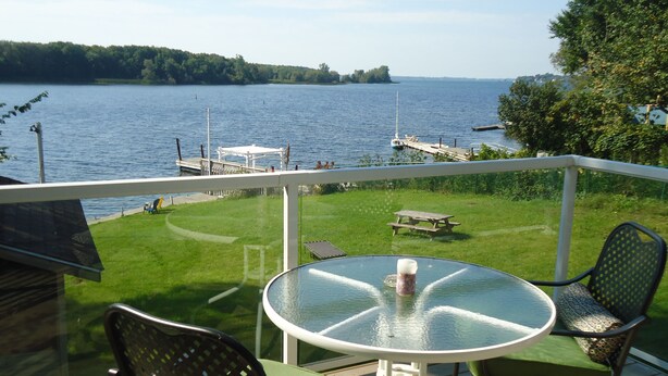 A balcony with a glass table, two chairs, and a view of a lake with boats and a dock. - Bayside Inn & Waterfront Suites (Kingston)
