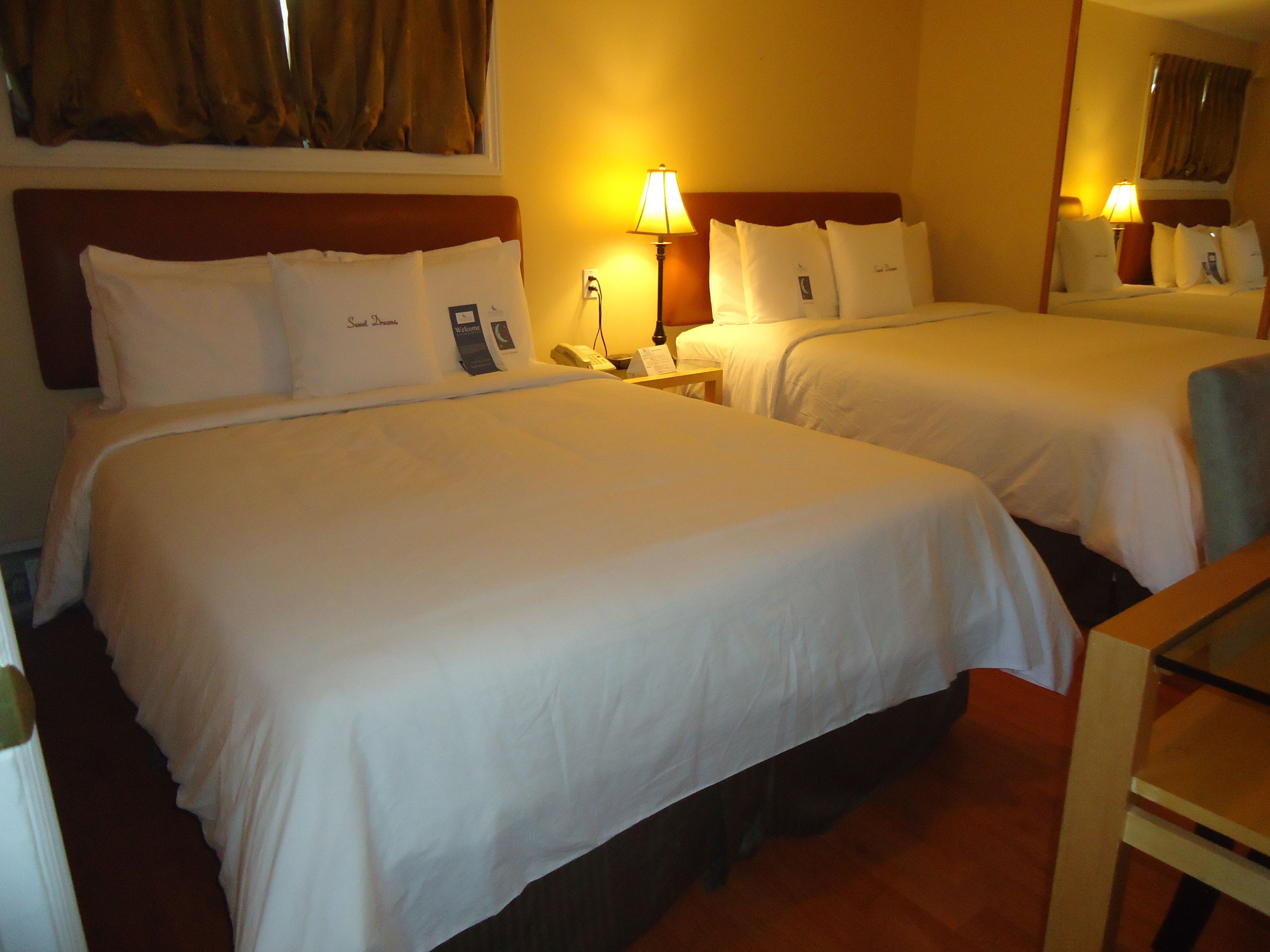 A hotel room with two single beds, each with white bedding and pillows, a bedside table with a lamp, and a mirror above the beds.