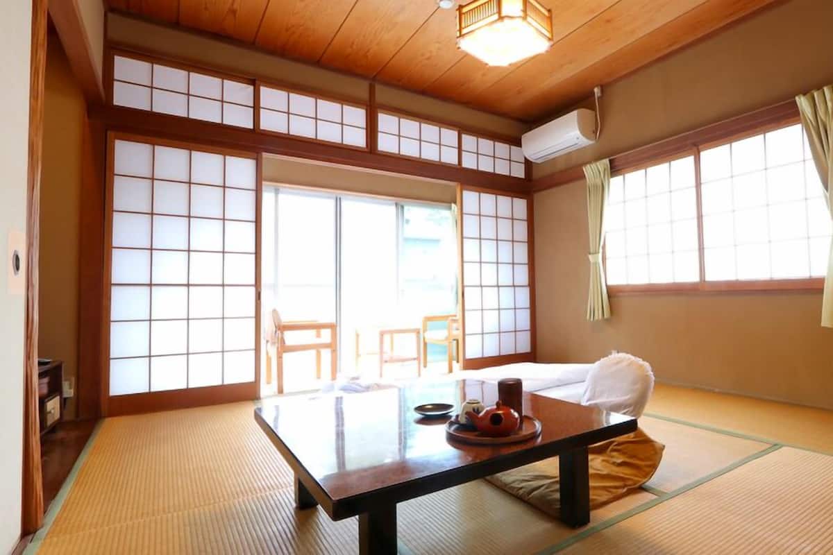 A traditional Japanese room with tatami flooring, a low table with a tea set, sliding doors, and a large window with curtains.