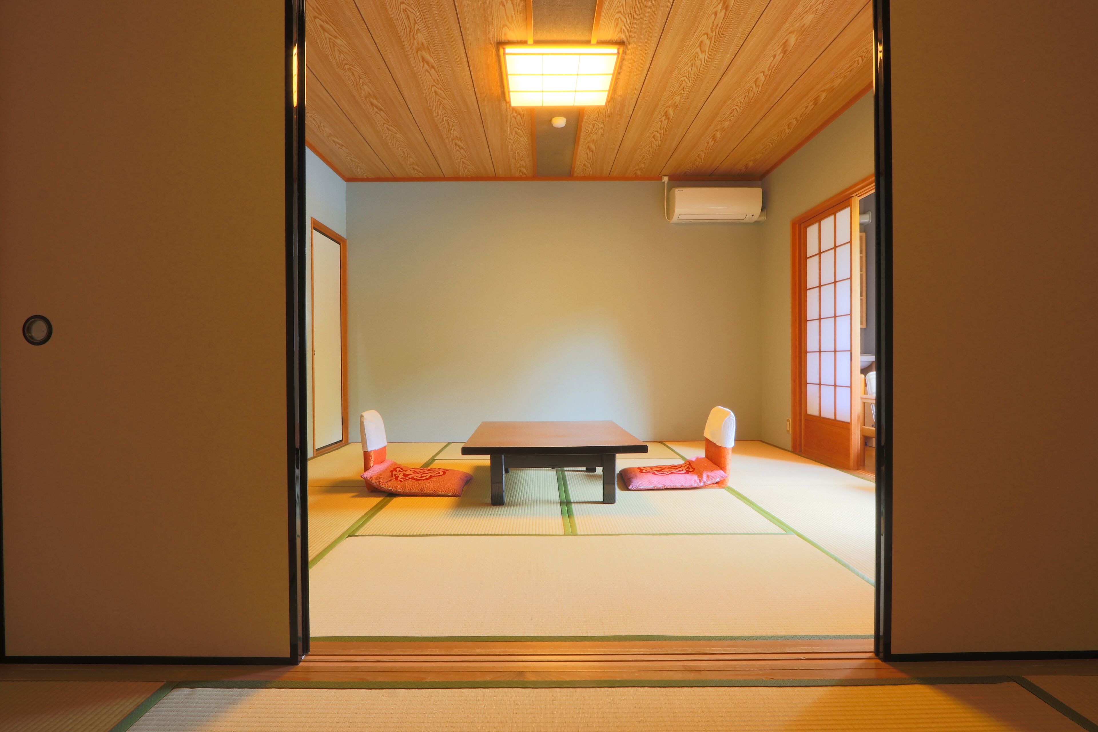 A tatami mat room with a low table and cushions, wooden ceiling, and sliding doors.