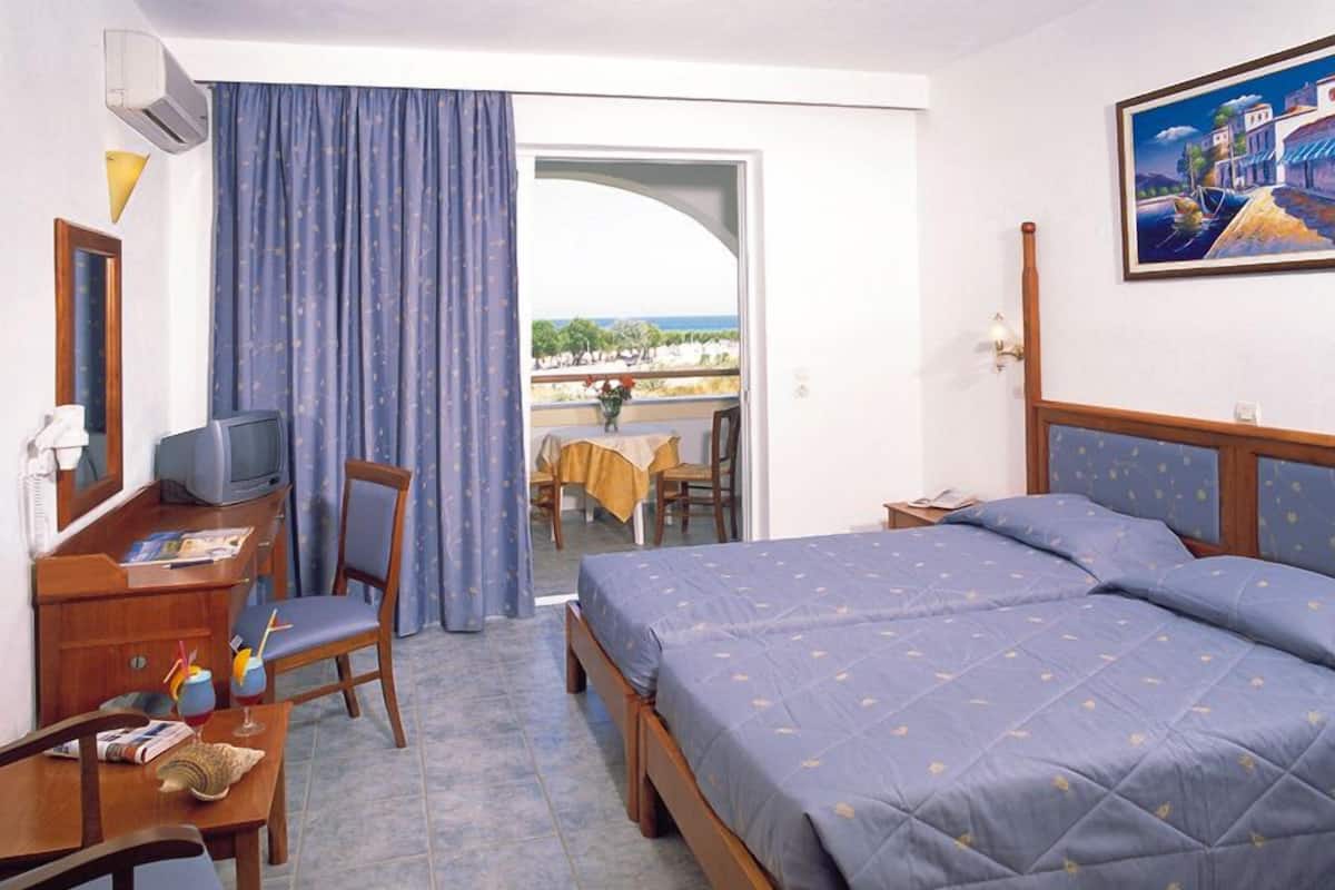 Une chambre d’hôtel avec un lit, un bureau avec une télévision, une chaise et une vue sur la mer par une porte ouverte.