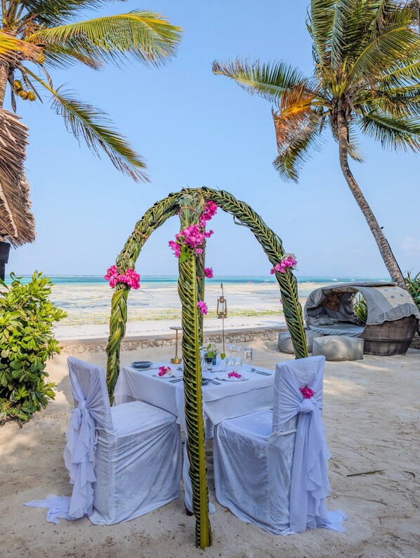 Une cérémonie de mariage organisée sur la plage, avec une arche de style tropical et des chaises blanches décorées de voilages.