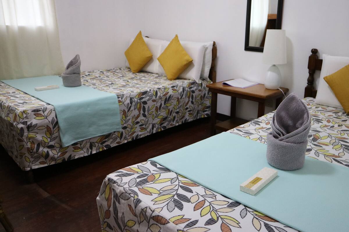 Une chambre d’hôtel avec deux lits simples, chacun recouvert d’une couvre-lit à motifs floraux et d’une couverture bleue. On y trouve une table de chevet en bois avec une lampe, une petite table avec un carnet, et une serviette sur le sol.