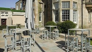 Un espace extérieur aménagé avec des tables et des chaises, un bâtiment en pierre doté de grandes fenêtres et un ciel dégagé.