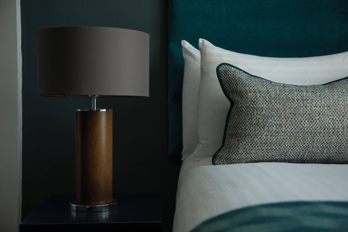 Un lit bien fait, avec du linge de lit blanc et un coussin turquoise, une lampe moderne à la base cylindrique et une table de chevet sombre.