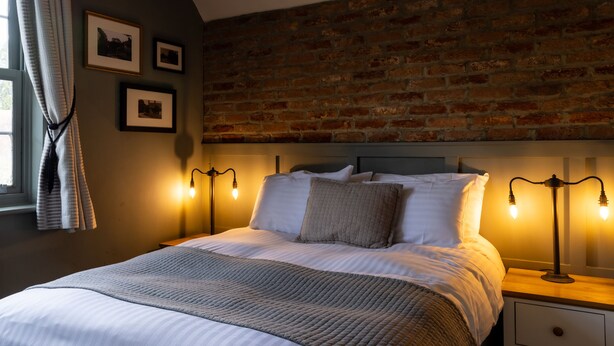 A bedroom with a brick wall, a bed with white bedding, two bedside lamps, and a window with curtains.