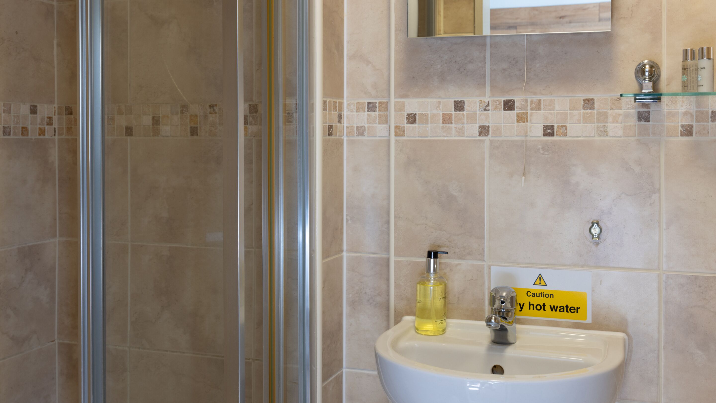 Un baño con cabina de ducha de vidrio, un lavamanos con una señal de precaución y una pared con azulejos.