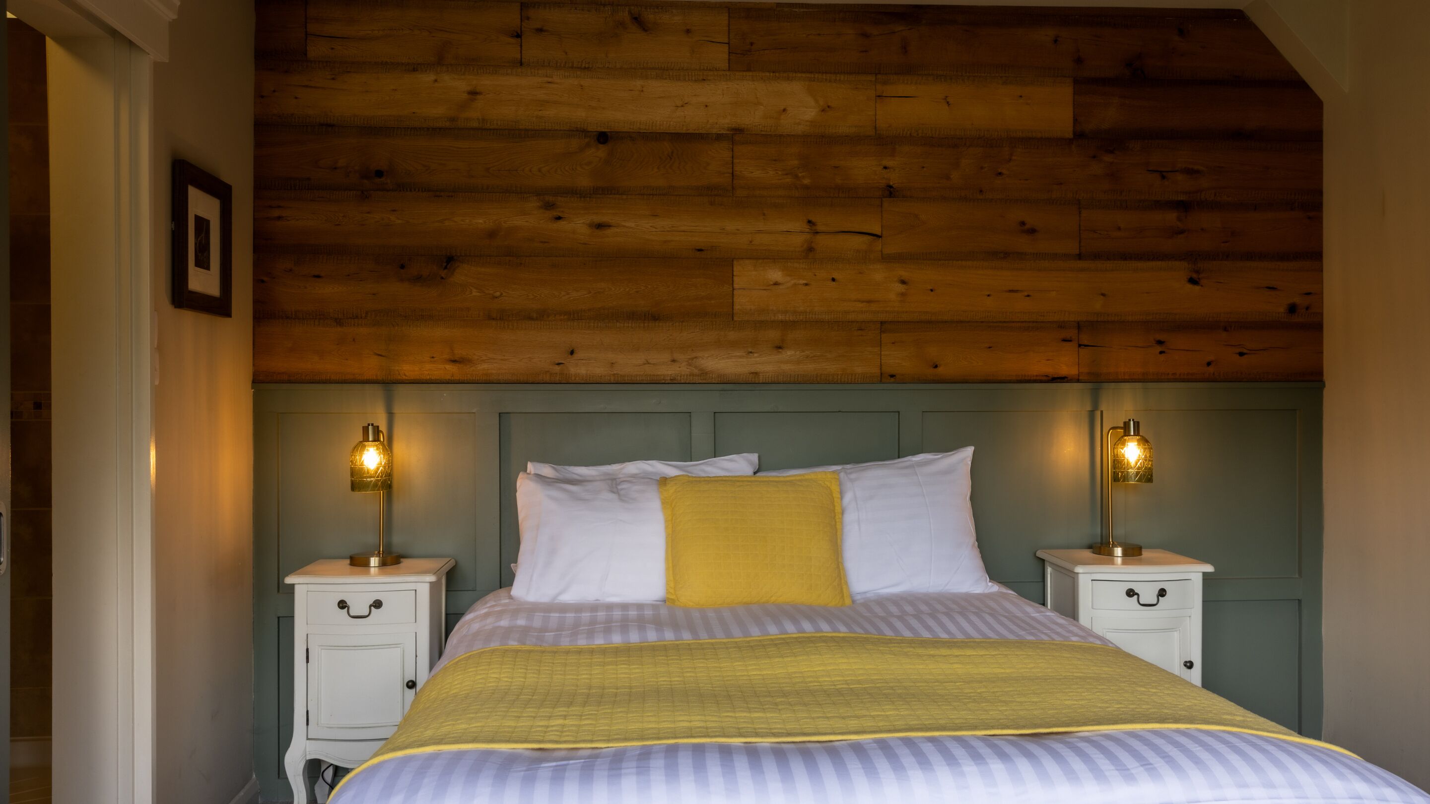 Un dormitorio con techo de madera, una cama con ropa de cama blanca y amarilla, dos mesitas de noche con lámparas y un cuadro enmarcado en la pared.