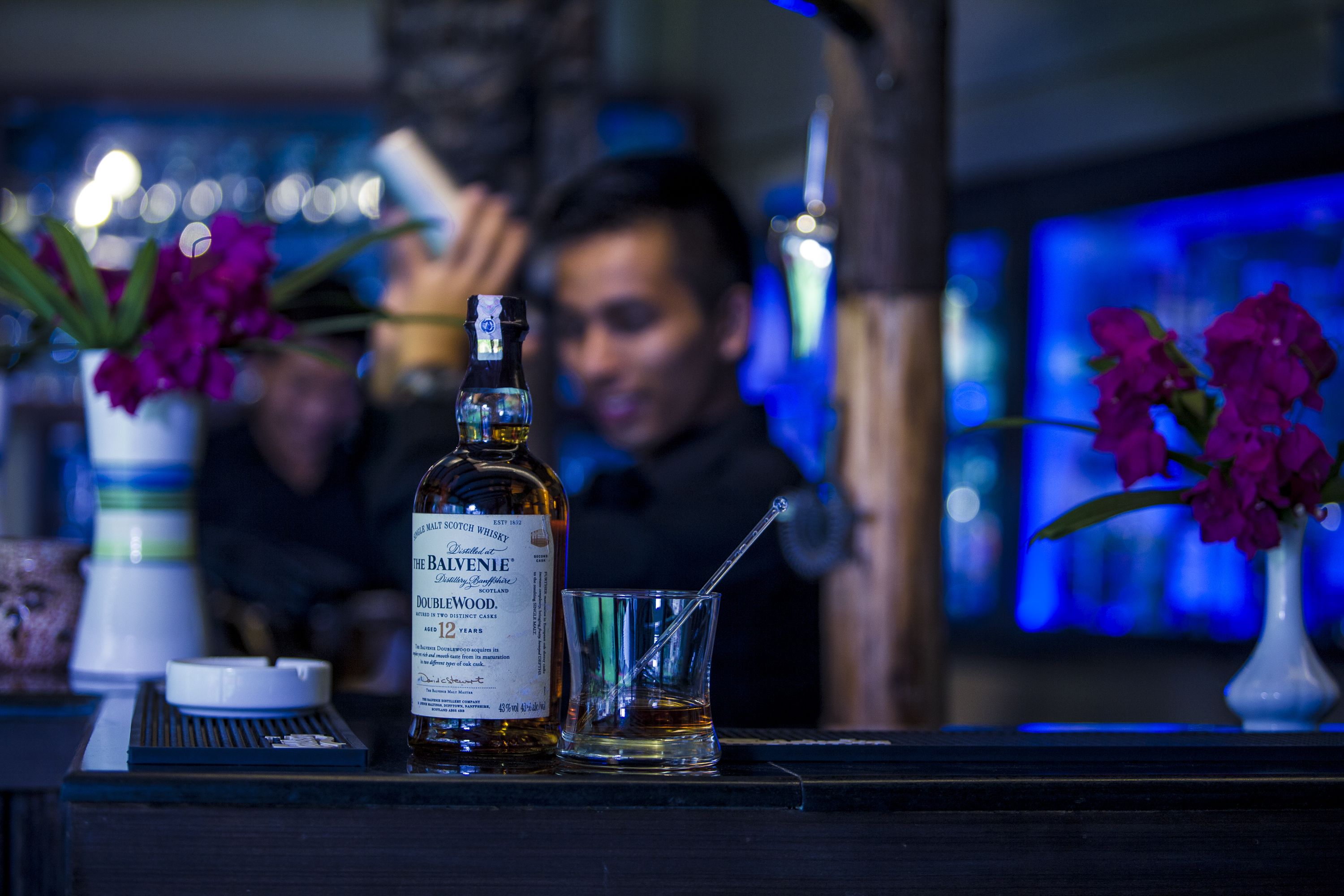Een fles The Balvenie Doublewood whisky, een glas met rietje en een kleine vaas met paarse bloemen op een barkruk.
