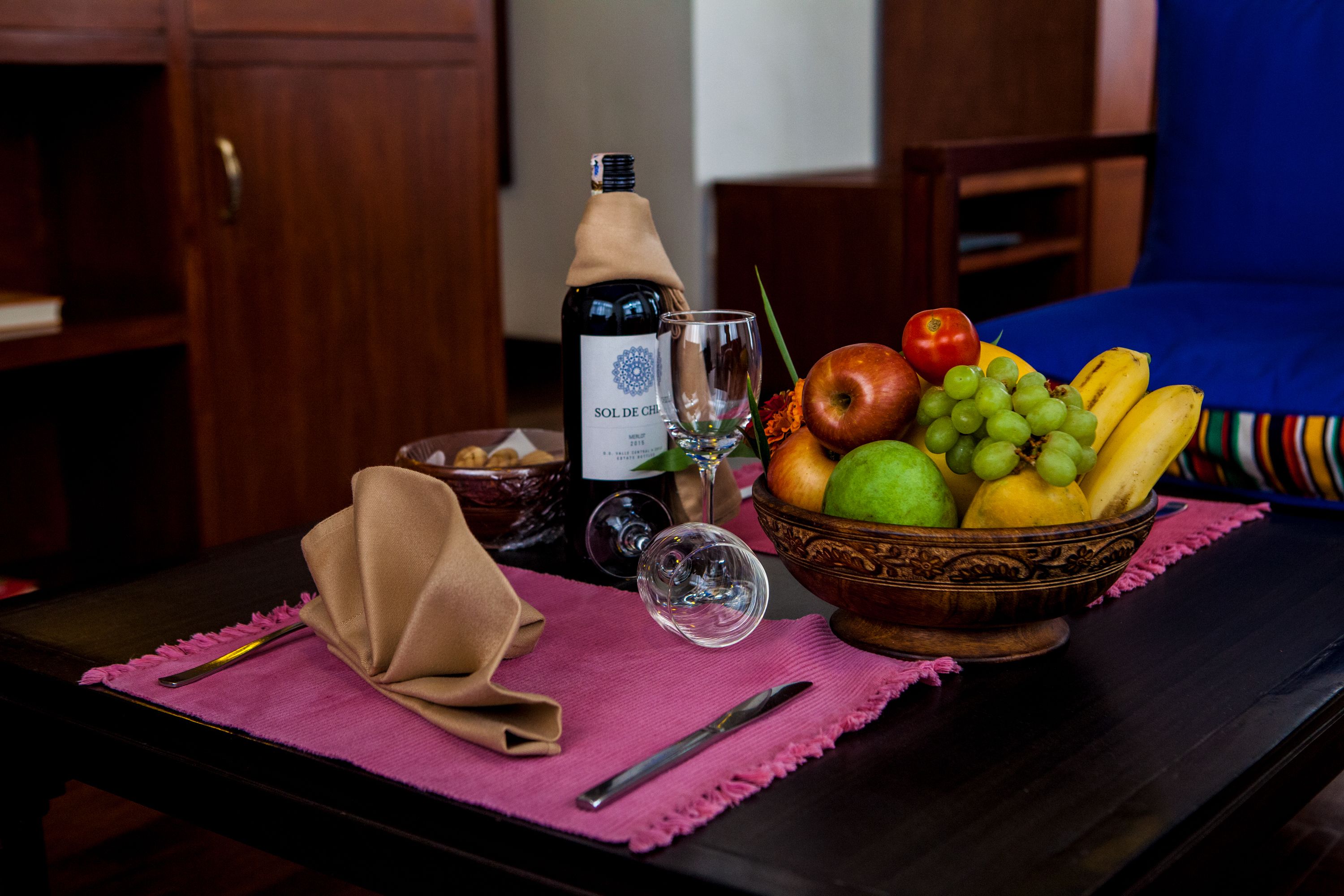 A table with a bottle of wine, a glass, a bowl of fruit, and a napkin on a pink placemat.