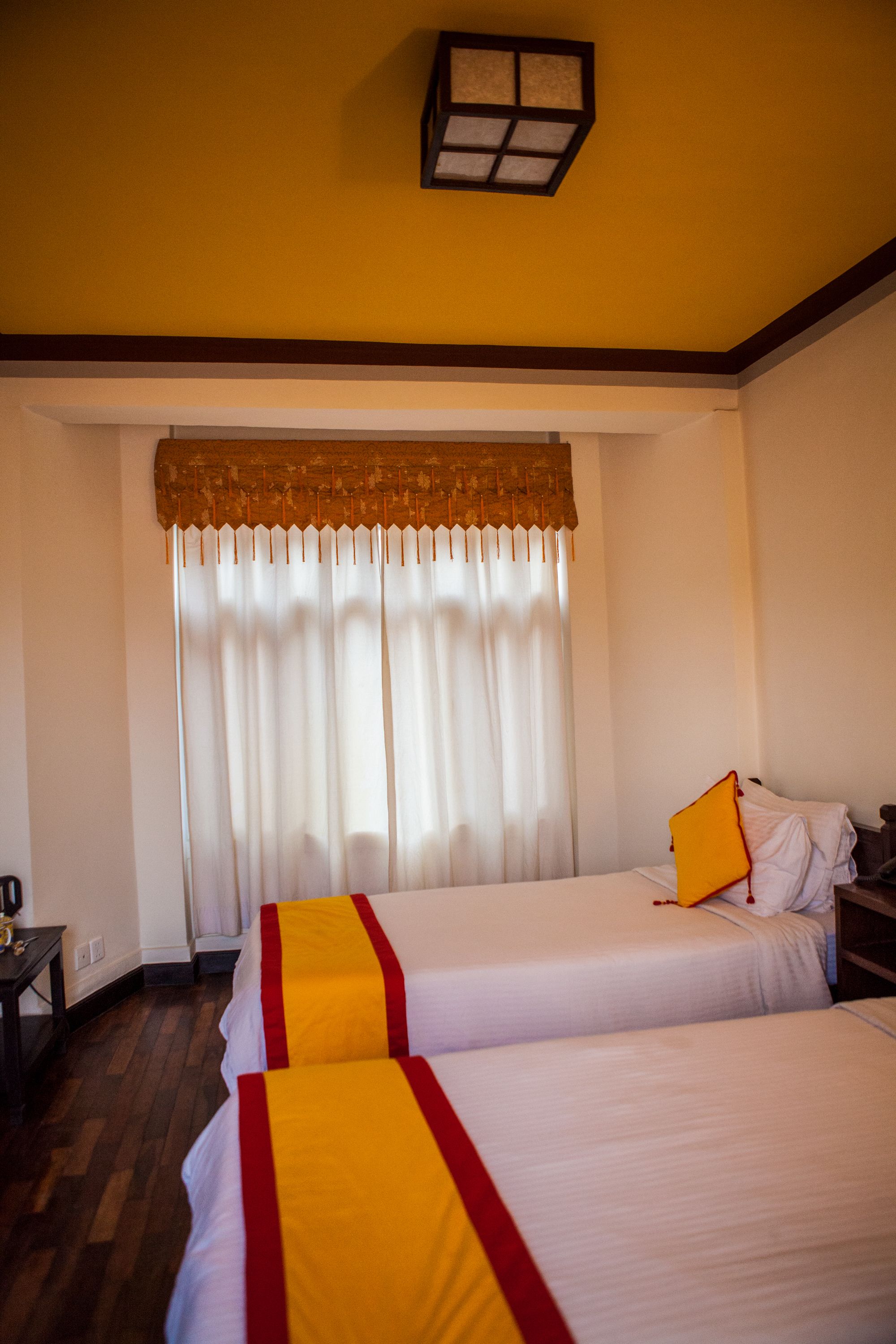 A hotel room with two beds, a wooden floor, a window with curtains, and a small table with a lamp.