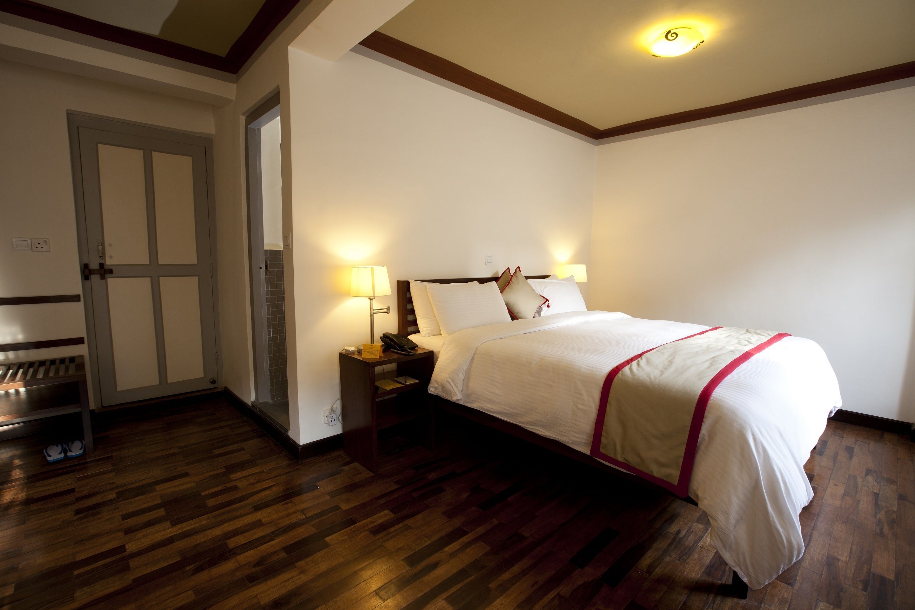 Una habitación de hotel con piso de madera, una cama con ropa blanca y un toque rojo, una mesita de noche con una lámpara y una puerta con un espejo.