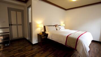 Una habitación de hotel con piso de madera, una cama con ropa blanca y un toque rojo, una mesita de noche con una lámpara y una puerta con un espejo.