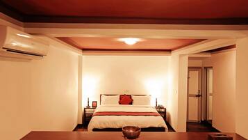 Quarto de hotel com cama, mesas de cabeceira, um criado-mudo, uma mesa com uma tigela e uma porta que dá para outro cômodo.