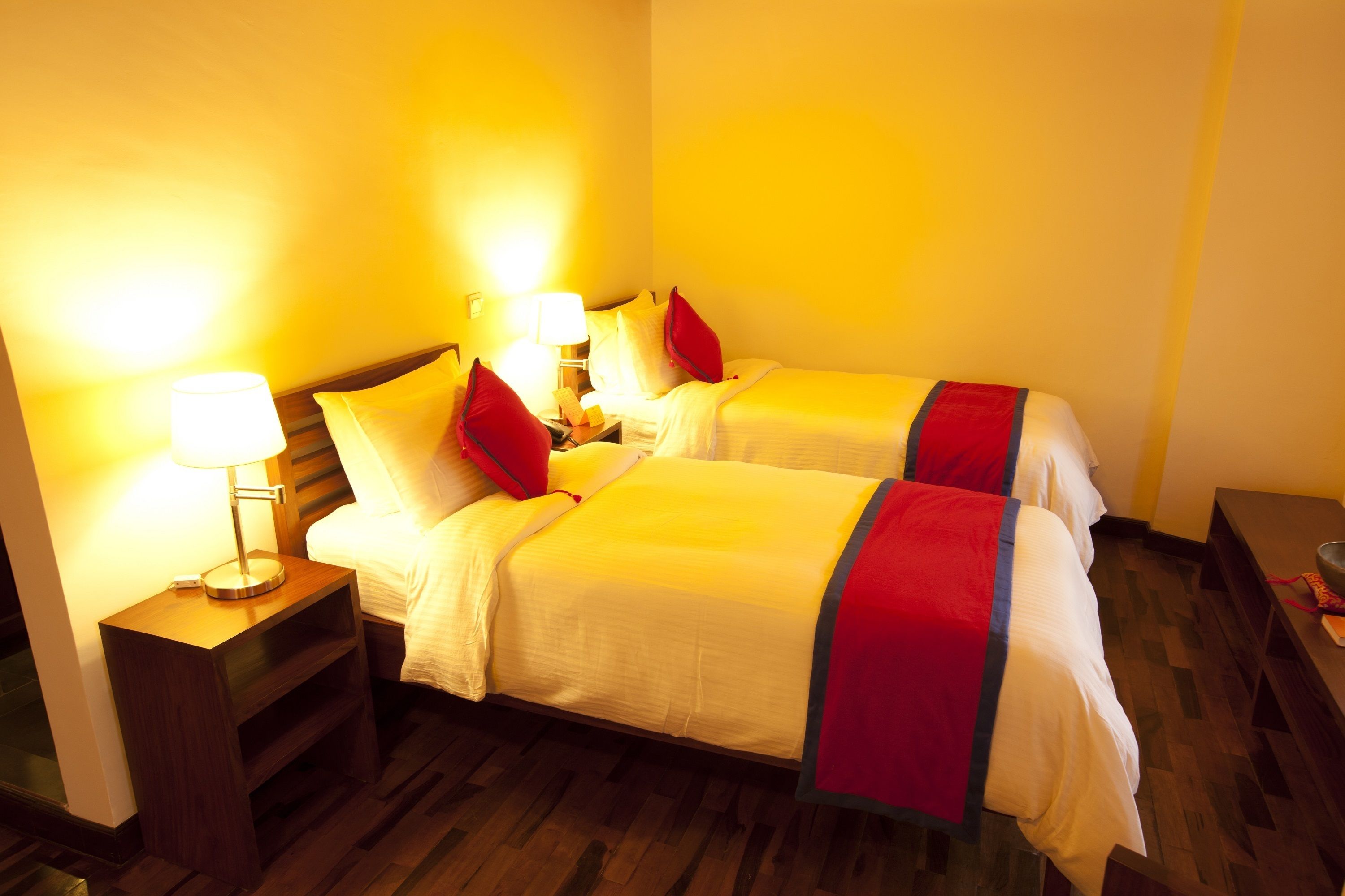 A hotel room with two beds, each with a red and white bedspread, a nightstand with a lamp, and wooden flooring.