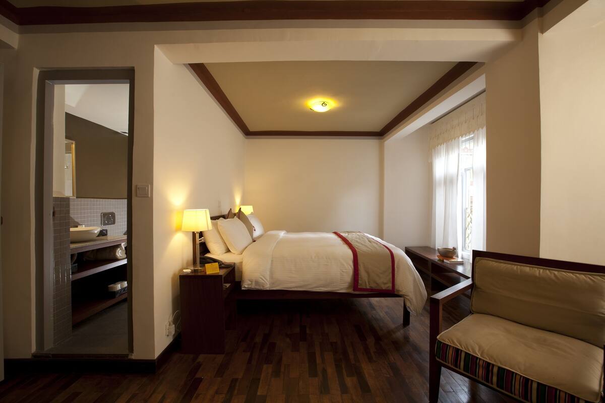 Een hotelkamer met een bed, nachtlampjes, een stoel, een nachtkastje en een raam met gordijnen.