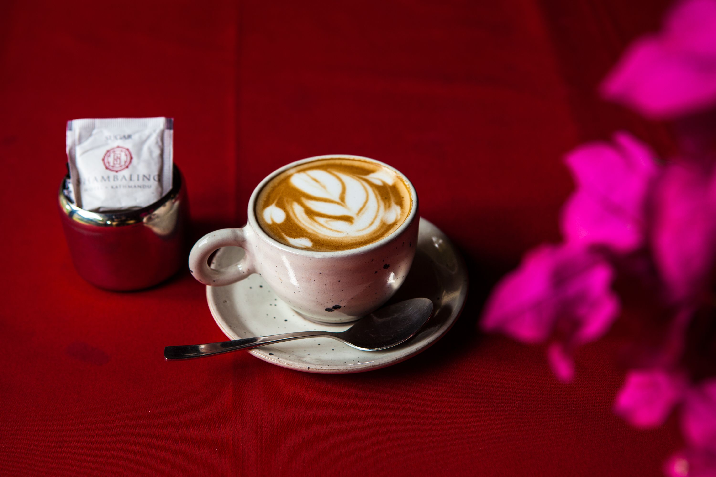 A cup of coffee with latte art on a saucer, a spoon, and a packet of sugar on a red table.