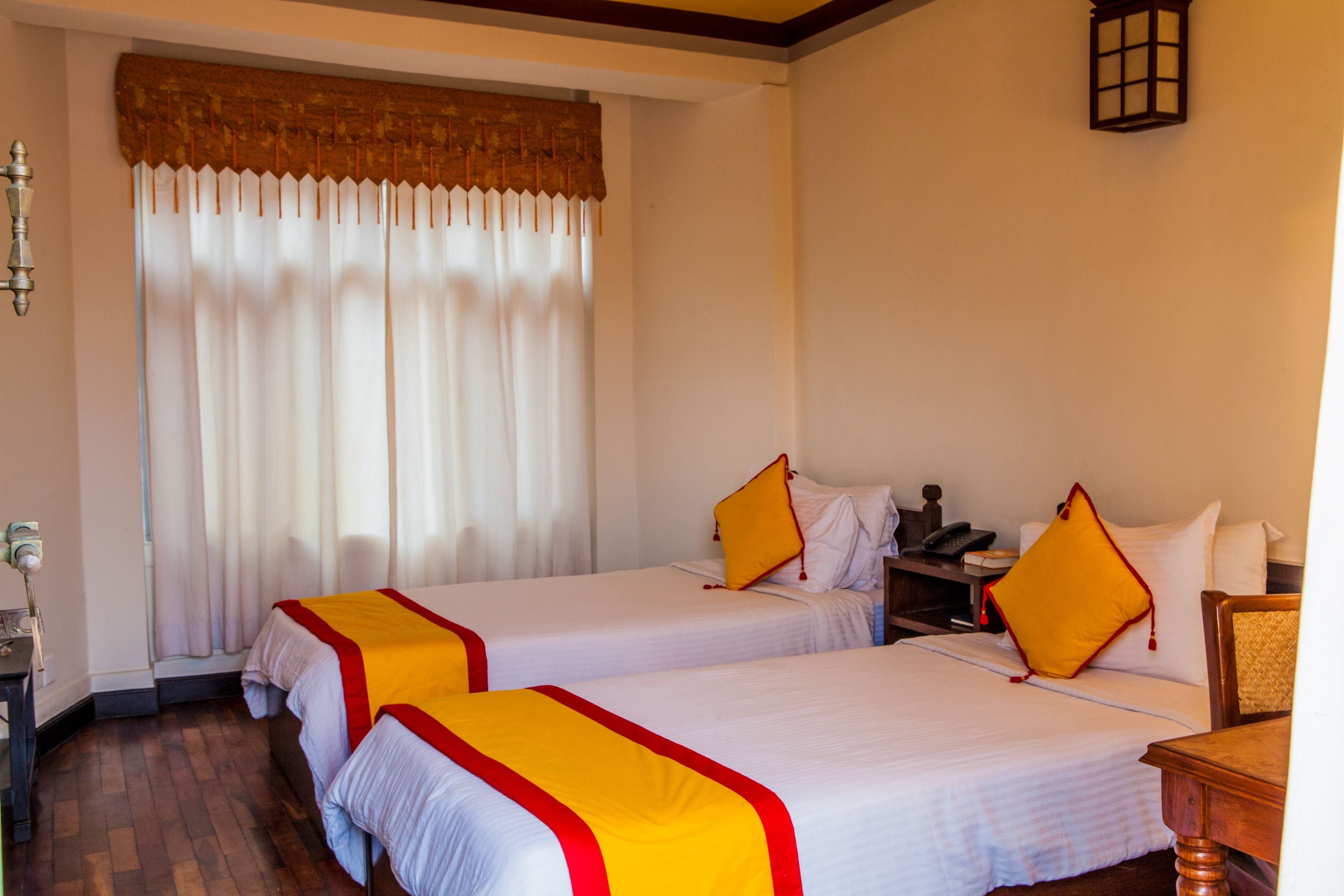 A hotel room with two beds, a wooden floor, a window with curtains, a nightstand, and a lamp.