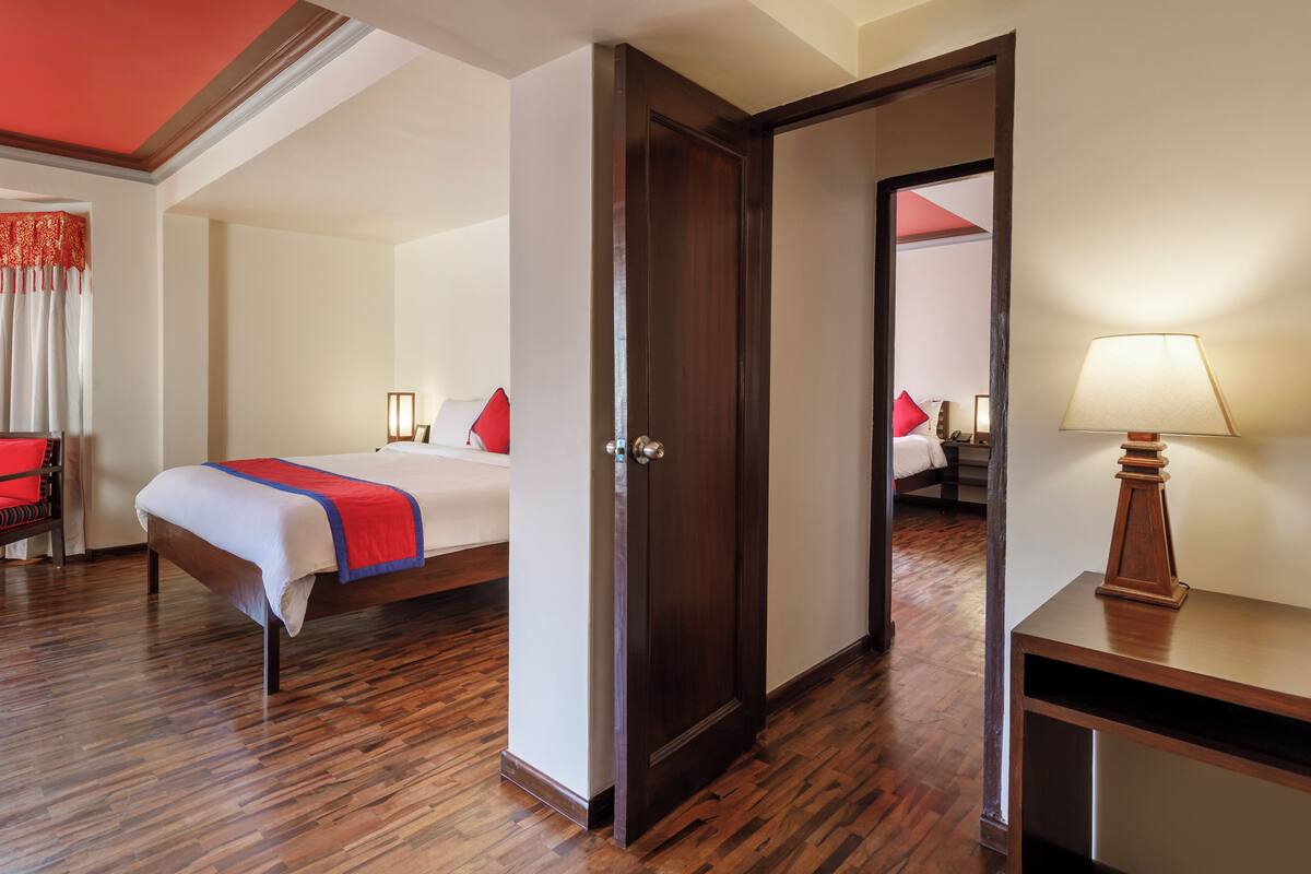 Een hotelkamer met een houten vloer, een bed met een rode en witte dekbedovertrek, een nachtkastje met een lamp, en een deur die naar een andere kamer leidt.