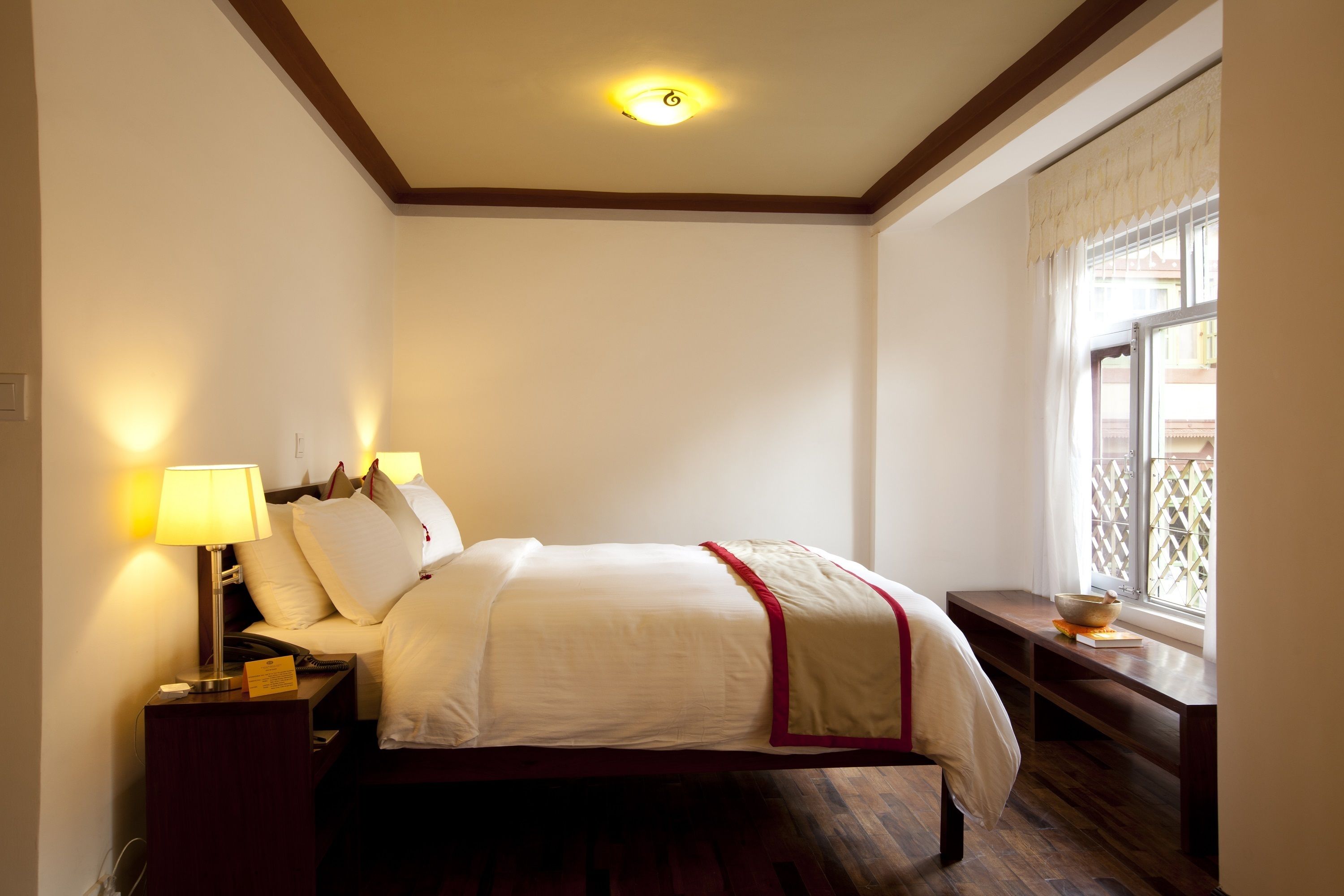 A neatly arranged hotel room with a bed, bedside lamps, a nightstand, a window with a view, and a wooden floor.