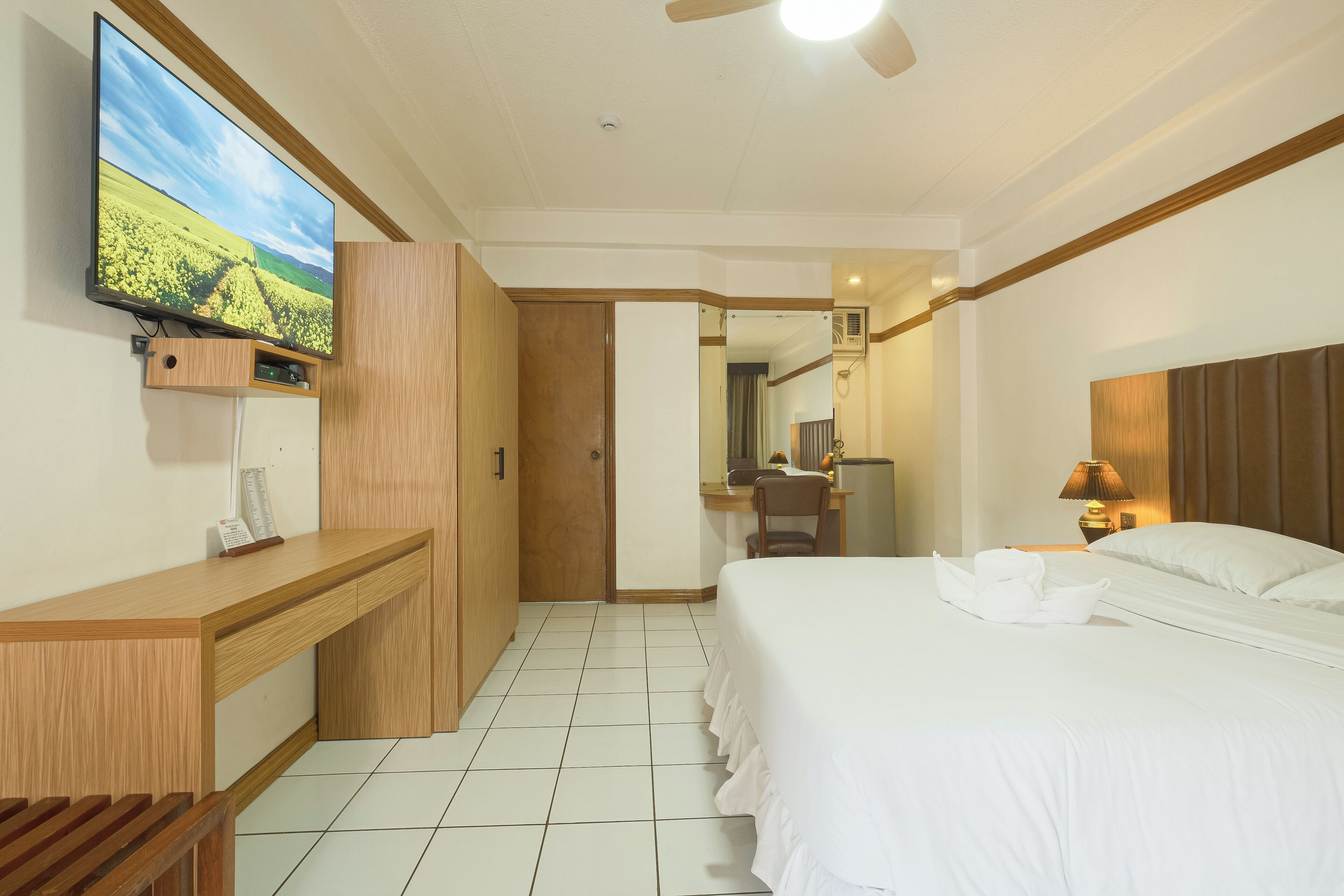 A hotel room with a bed, a desk, a television mounted on the wall, and a ceiling fan.