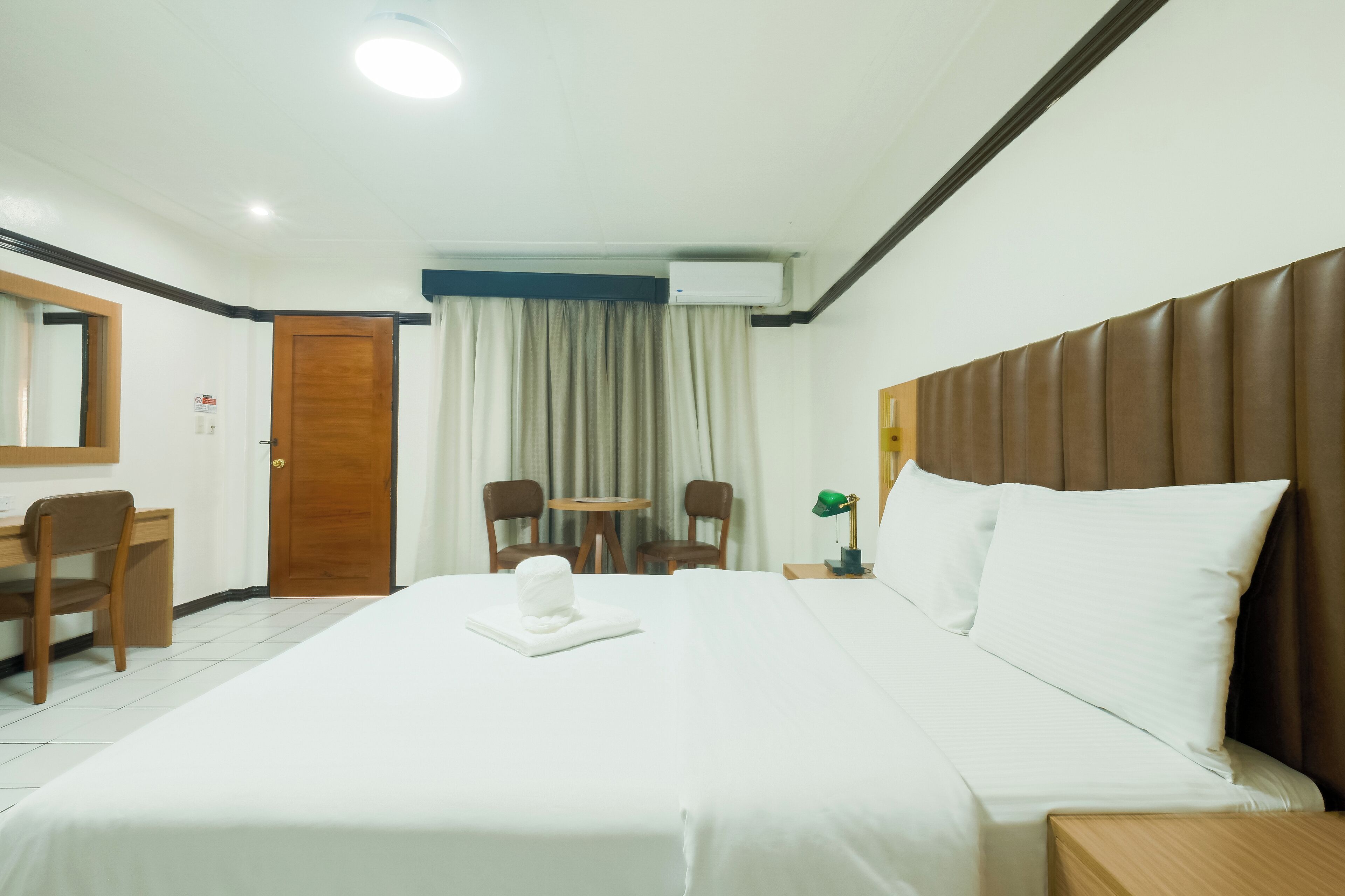 A hotel room with a large bed, a desk, two chairs, a small table, and a window with curtains.