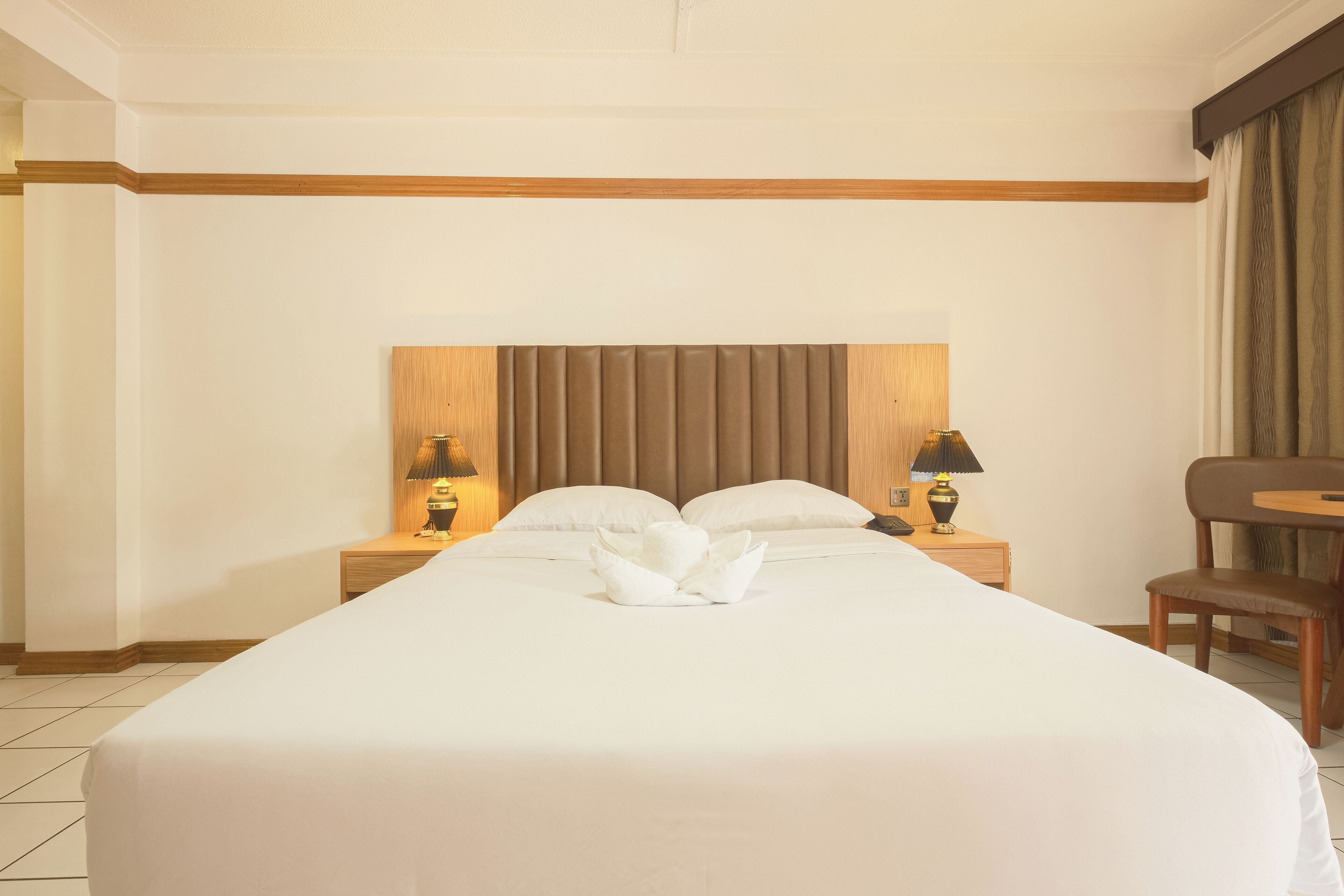 A hotel room with a large bed, two bedside tables with lamps, a wooden headboard, and a chair by the window.