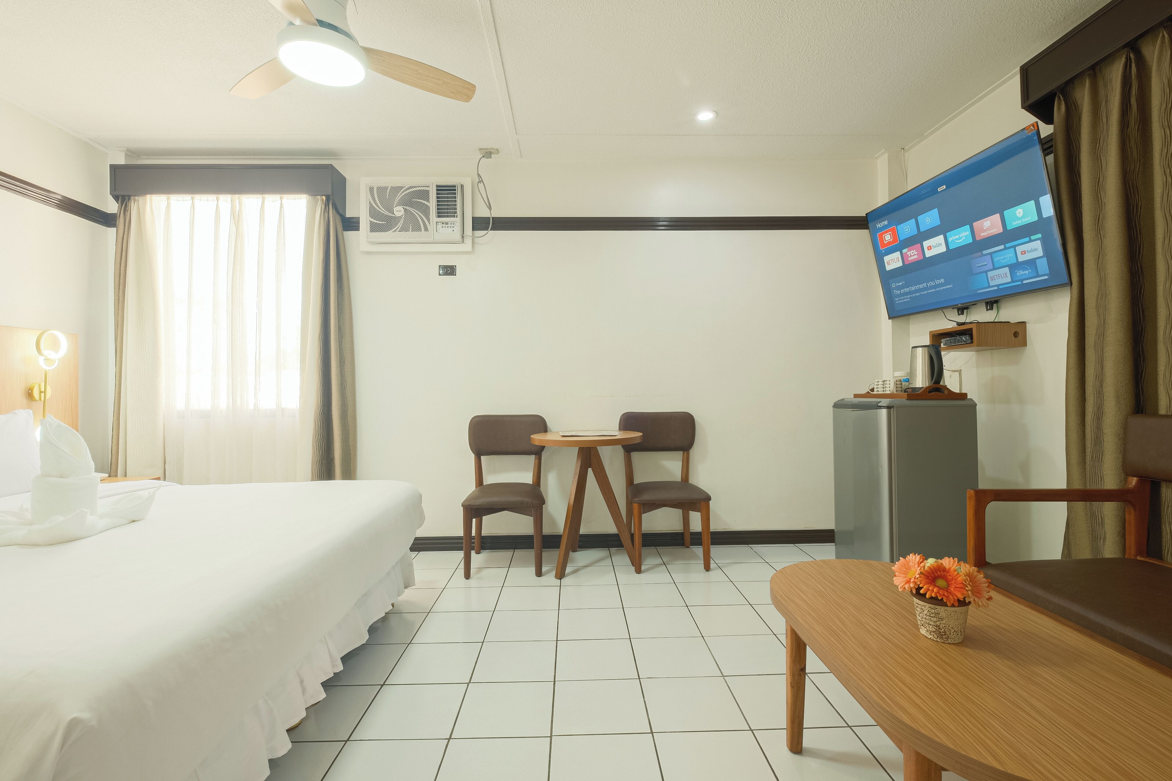 A hotel room with a bed, two chairs, a small table, a TV, and a desk with a keyboard.