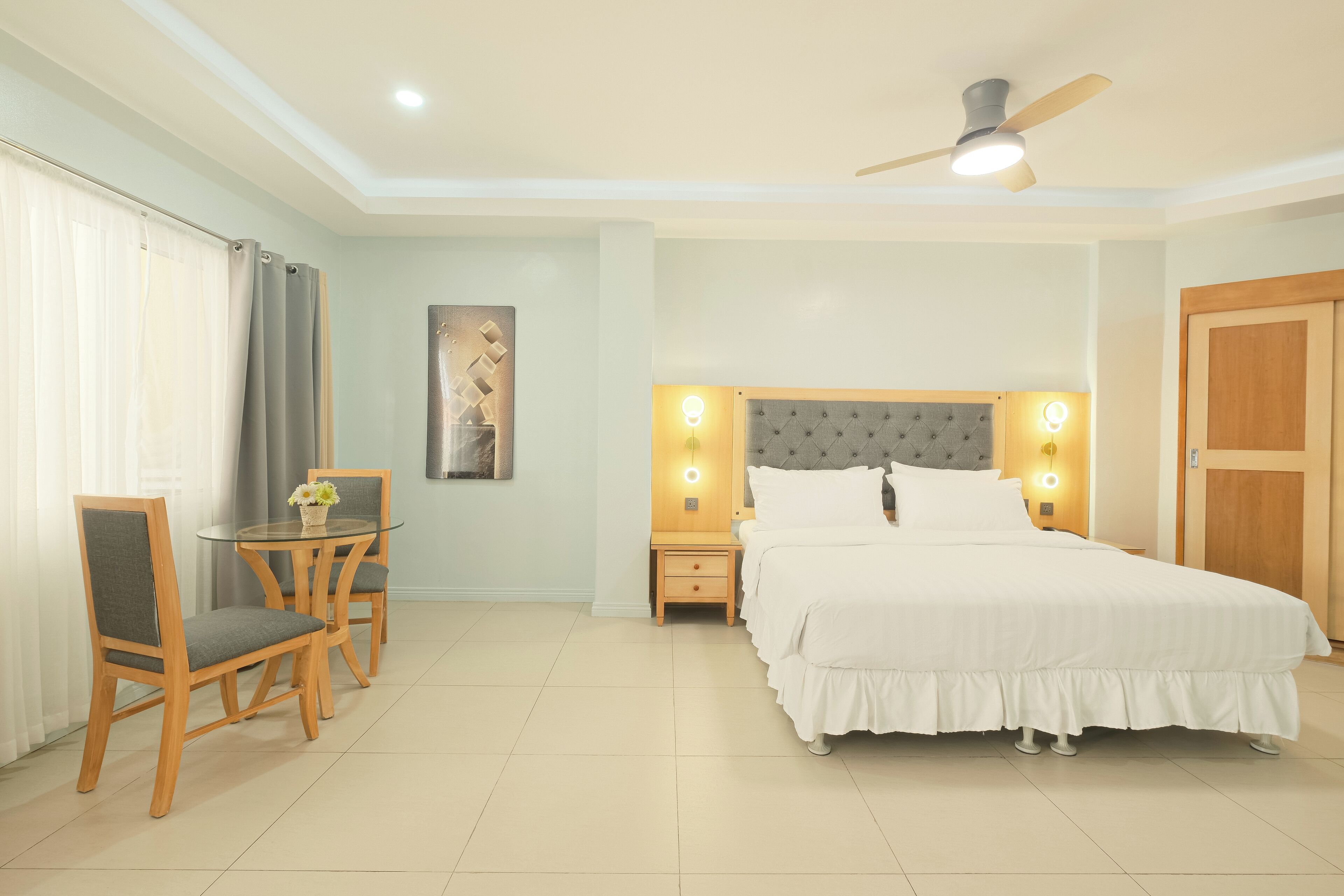 A bedroom with a bed, a desk with chairs, a nightstand with a lamp, and a ceiling fan.