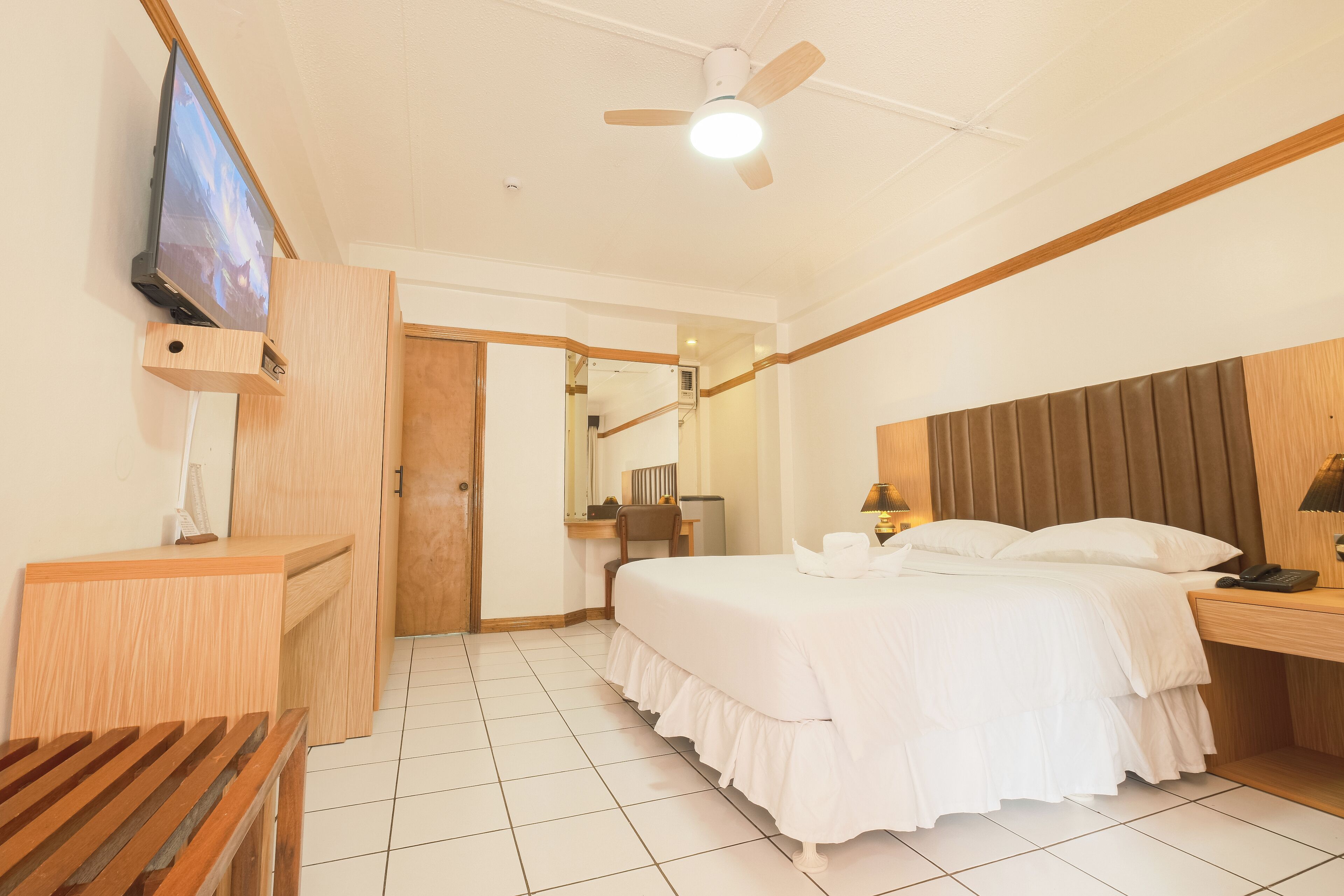 A hotel room with a bed, bedside table, TV mounted on a wooden panel, a ceiling fan, and a wooden bench.