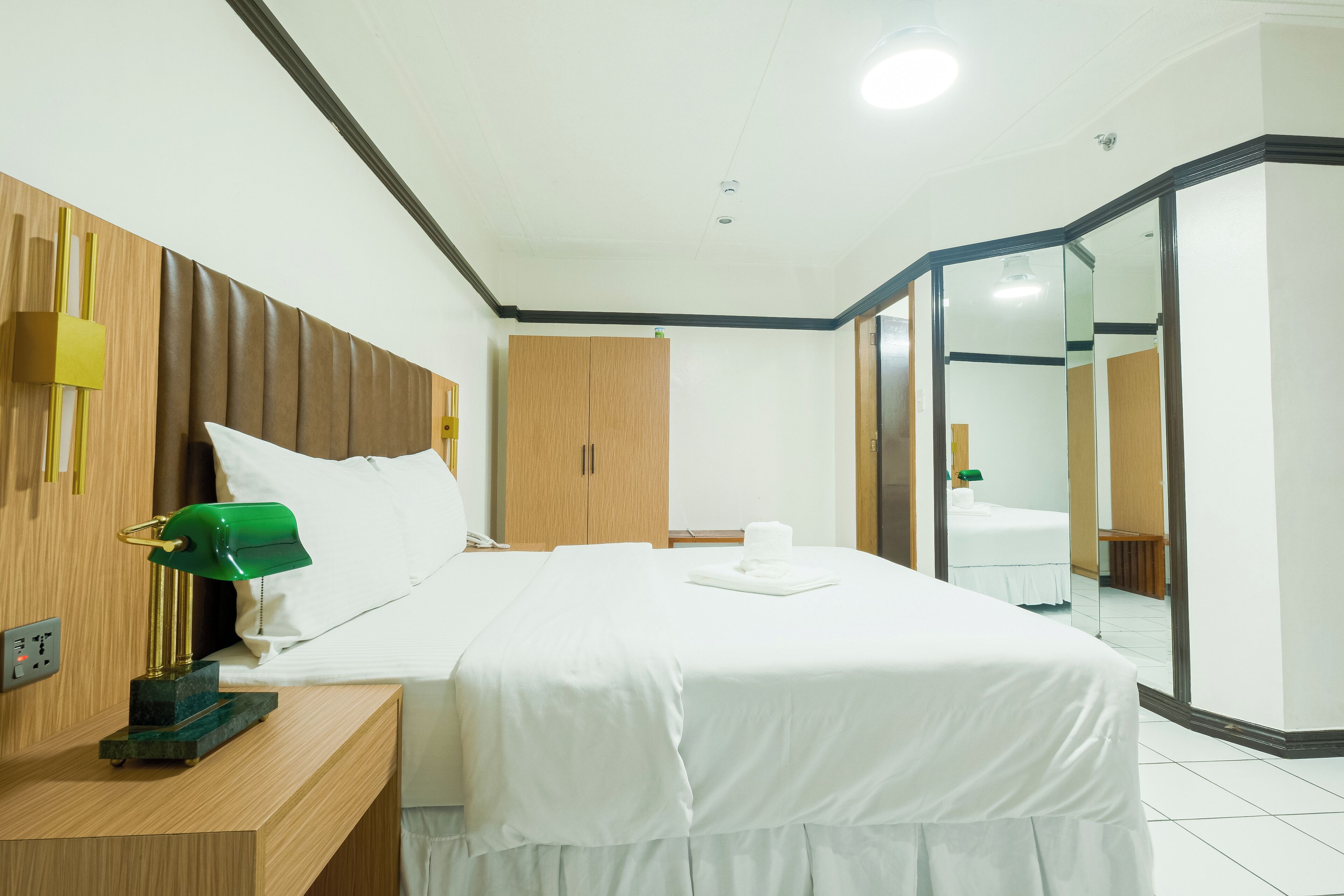 Ein Hotelzimmer mit Bett, Holz-Kopfteil, Nachttisch mit Lampe, Kleiderschrank und Spiegel.