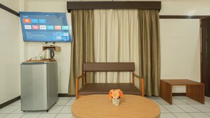 Ein Hotelzimmer mit Flachbildfernseher, der Streaming-Dienste anzeigt, einem Kühlschrank, einer hölzernen Bank, einem kleinen Tisch mit Blumenvase und einem Vorhang.