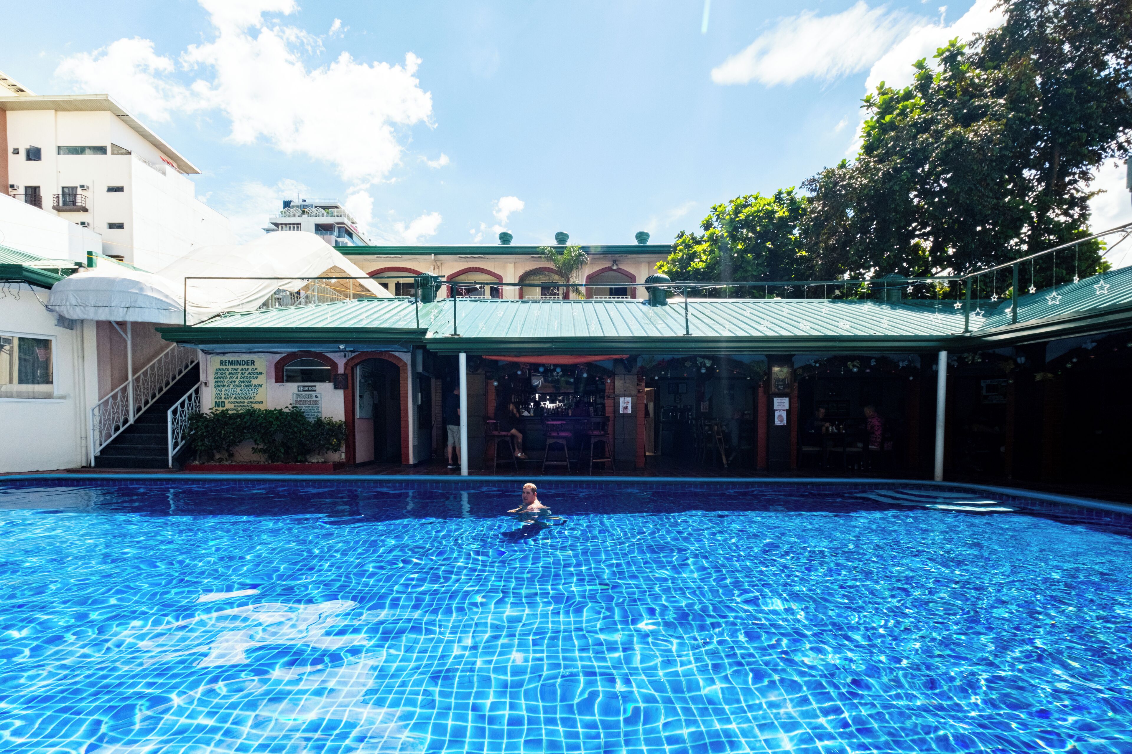 A swimming pool with a person in the water, surrounded by buildings with a green roof and a bar area.
