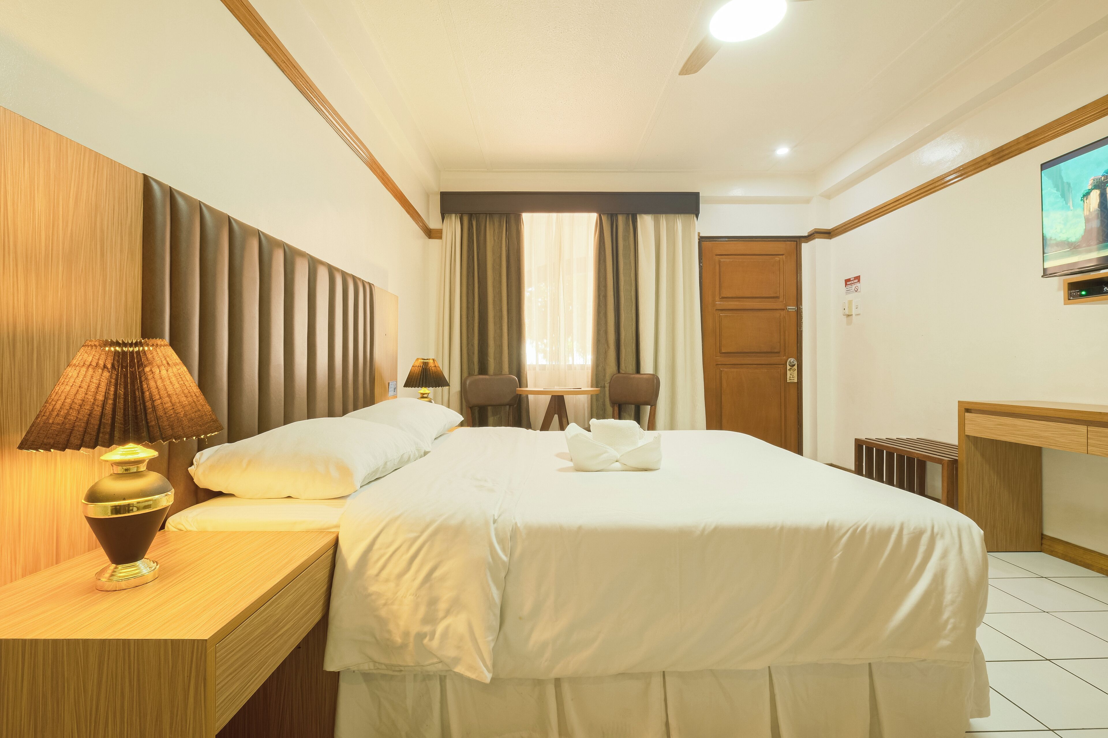 Habitación de hotel con cama, lámpara de noche, cabecera de madera y televisión en la pared.