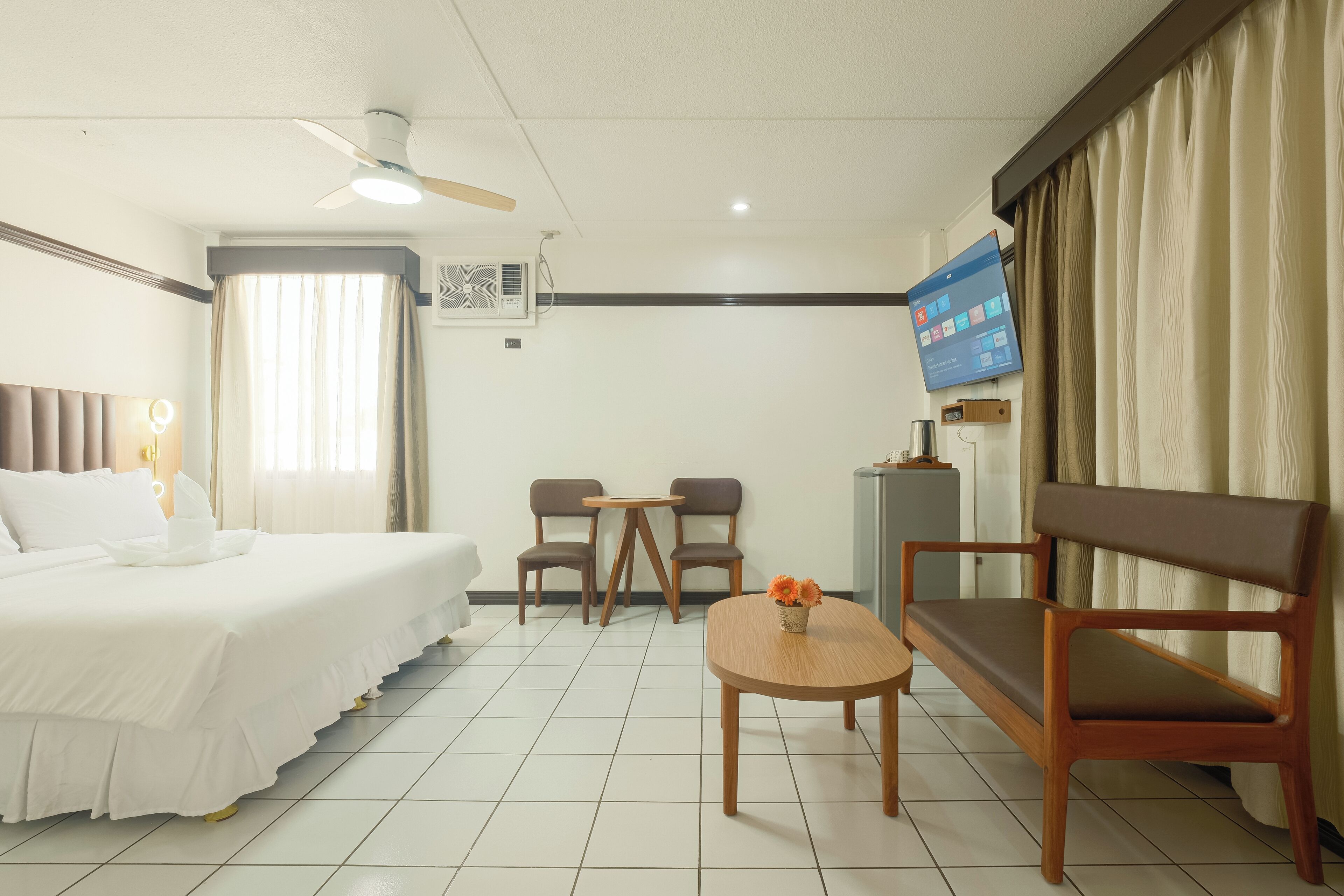 A hotel room with a bed, a desk, two chairs, a TV, and a ceiling fan.