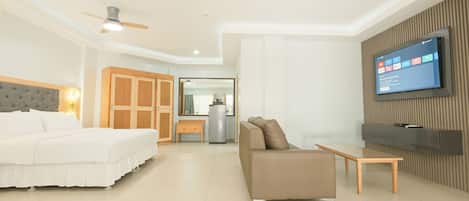 A modern hotel room with a bed, sofa, TV, and a kitchenette in the background.