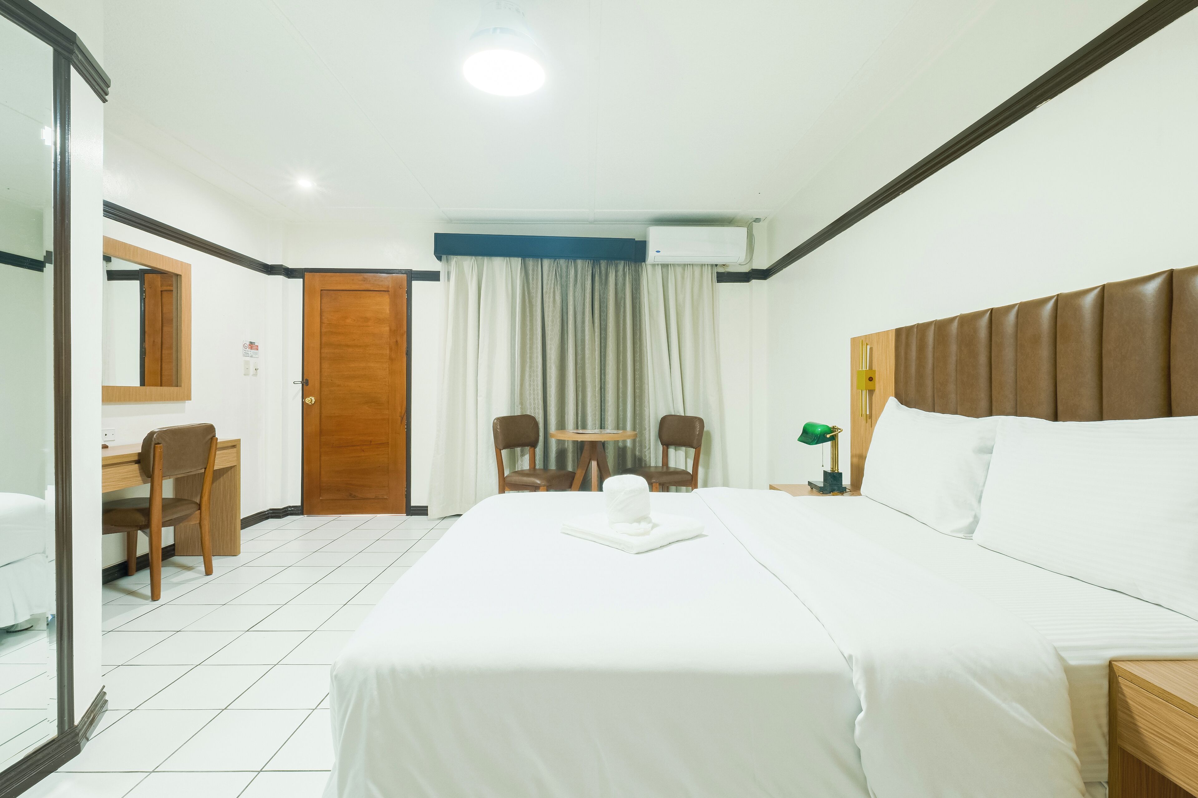 A hotel room with a bed, a desk, two chairs, a small table, and an air conditioning unit.
